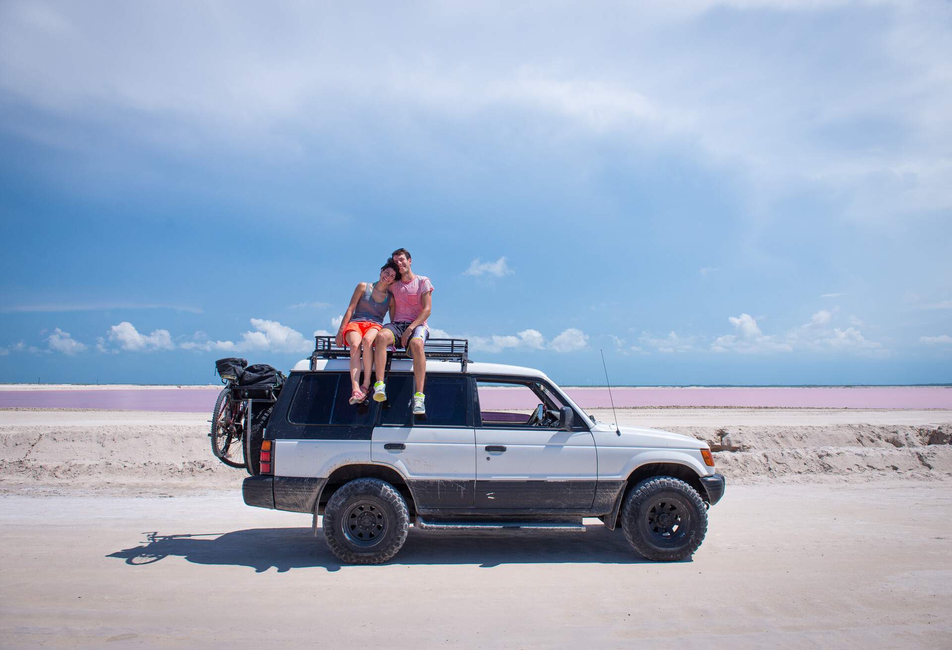 Couple sitting at the top of their car. They are hugging and looking to the camera. Girl leans on guy's shoulder. Pink water and white sand background. Travel concept. ; Shutterstock ID 1214050318; Purpose: Product; Brand (KAYAK, Momondo, Any): Any