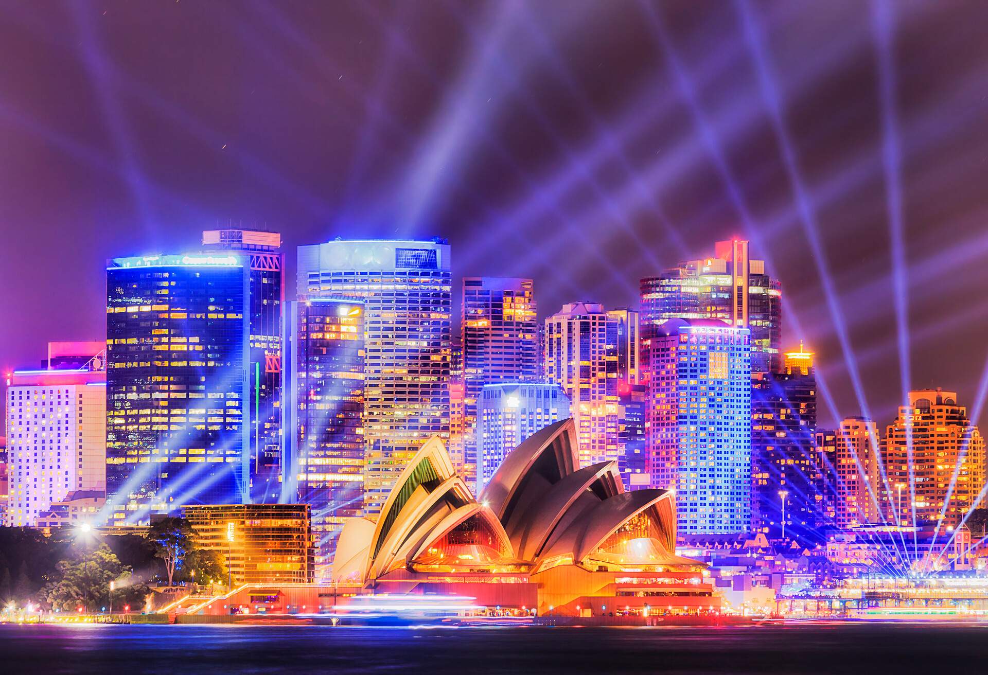 Wall of high-rise office towers on Sydney city waterfront of Circular quay during annual light show with bright blue beams in dark sky over major city landmarks across the harbour.