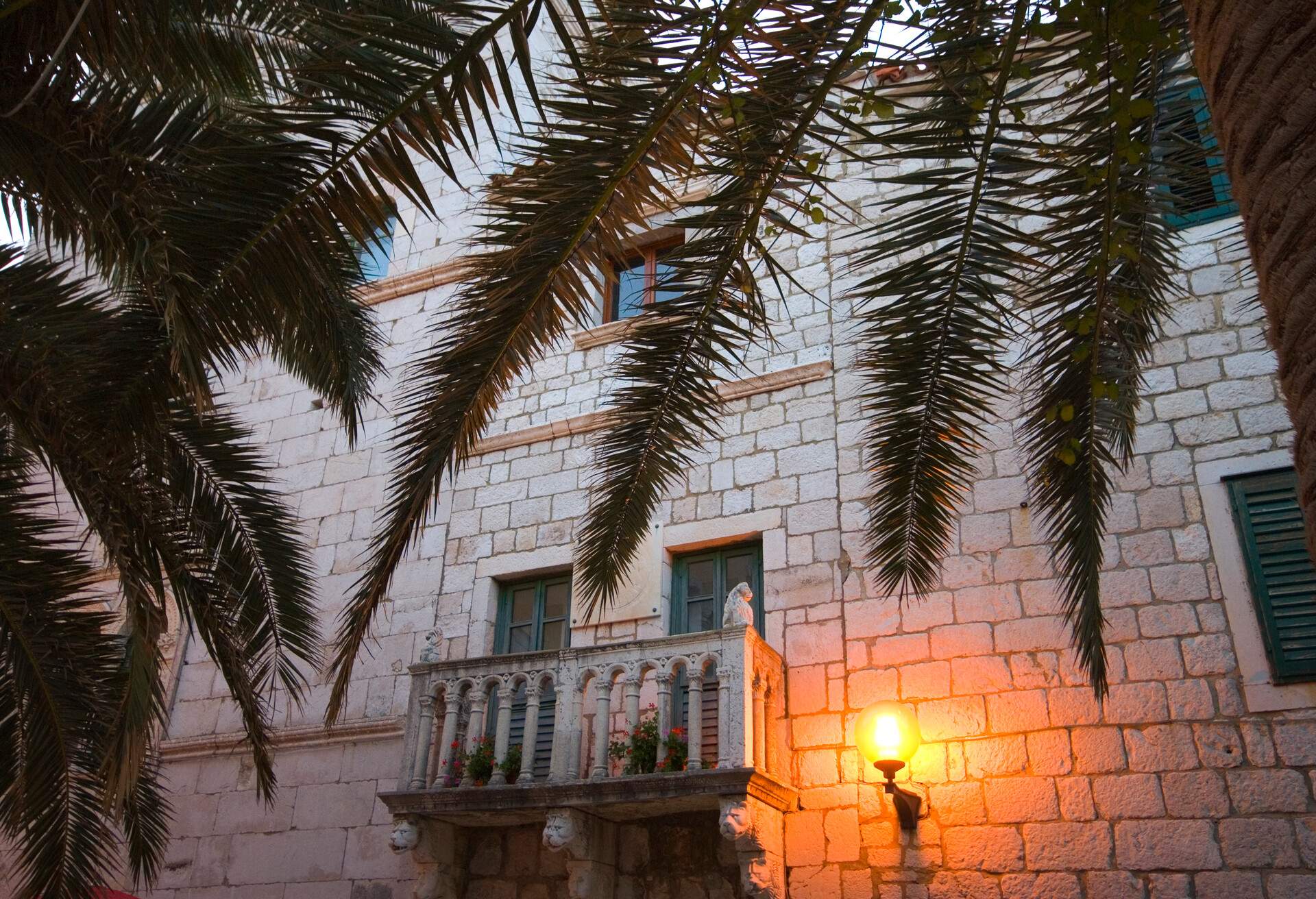 Facade with balcony through the palms of Municipium Arbae square.