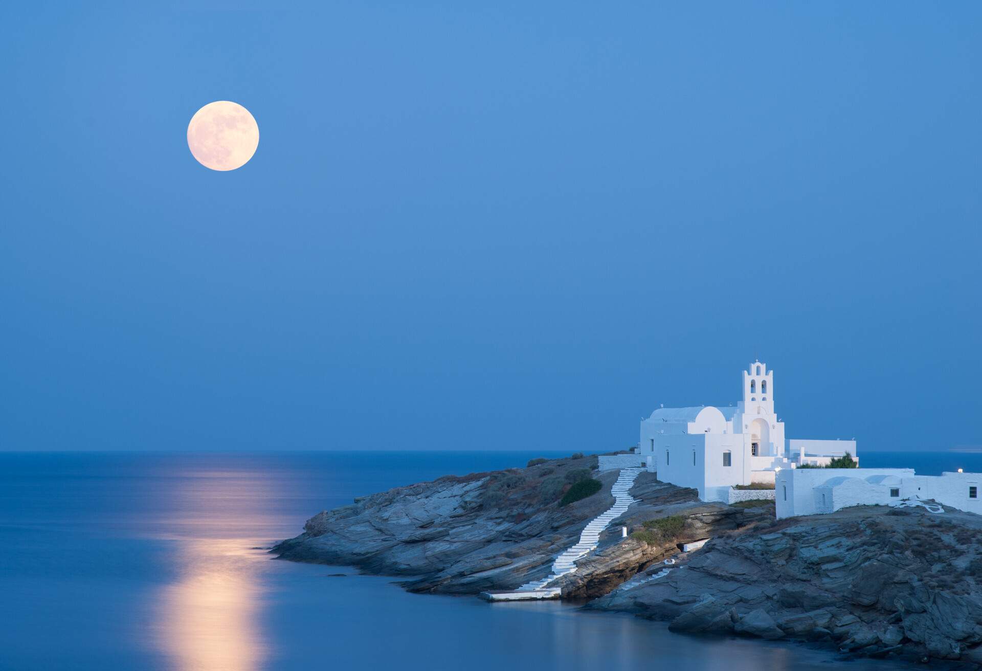 A summer picture of the island Sifnos with the full moon and its reflections on the Aegean Sea. The famous church of Panagia Chrysopigi on the foreground.