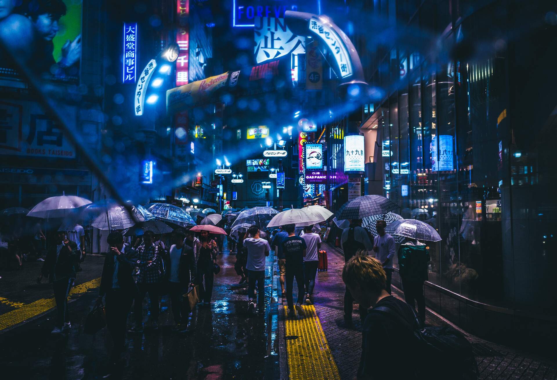 Amidst a rain-soaked night, people scurry along the streets, clutching umbrellas, as the brightly-lit signs and advertisements of the cityscape shimmer and reflect in the wet pavement.