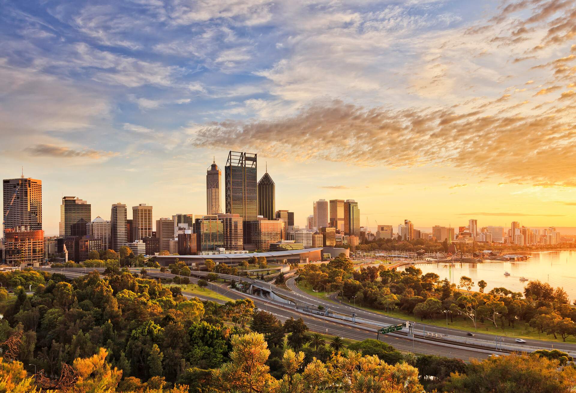 Gold warm sun light litting CBD of Perth city as seen from Kings park with green trees and highway entering the city.