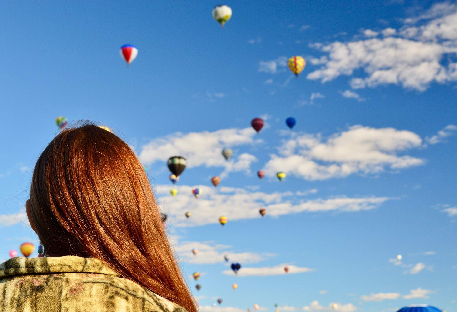 An individual looks at the colourful floating hot air balloons in the cloudy blue sky.