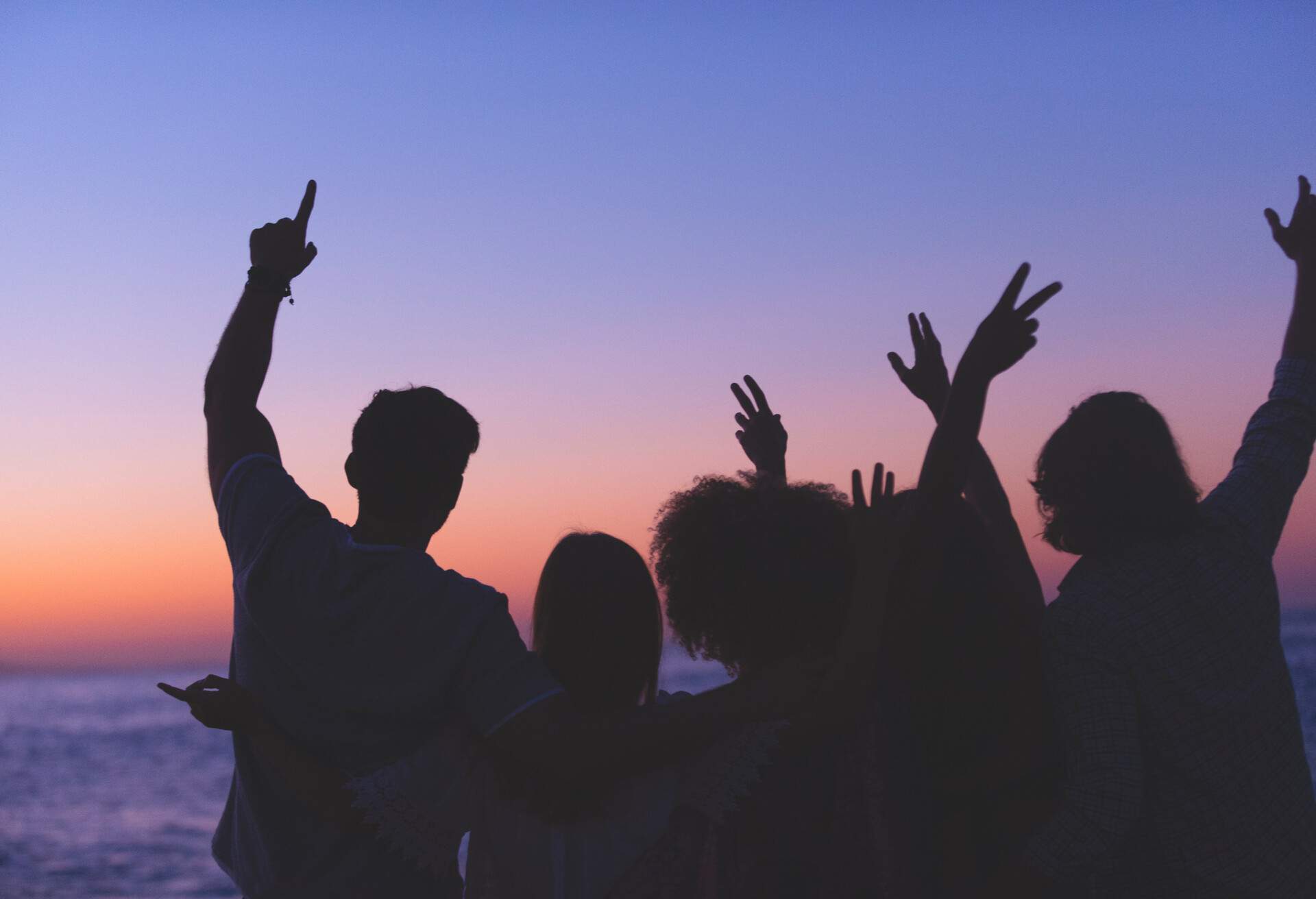 Group of people partying on the beach at sunset or sunrise. They have their arms raised in celebration. Backlit silhouette with copy space