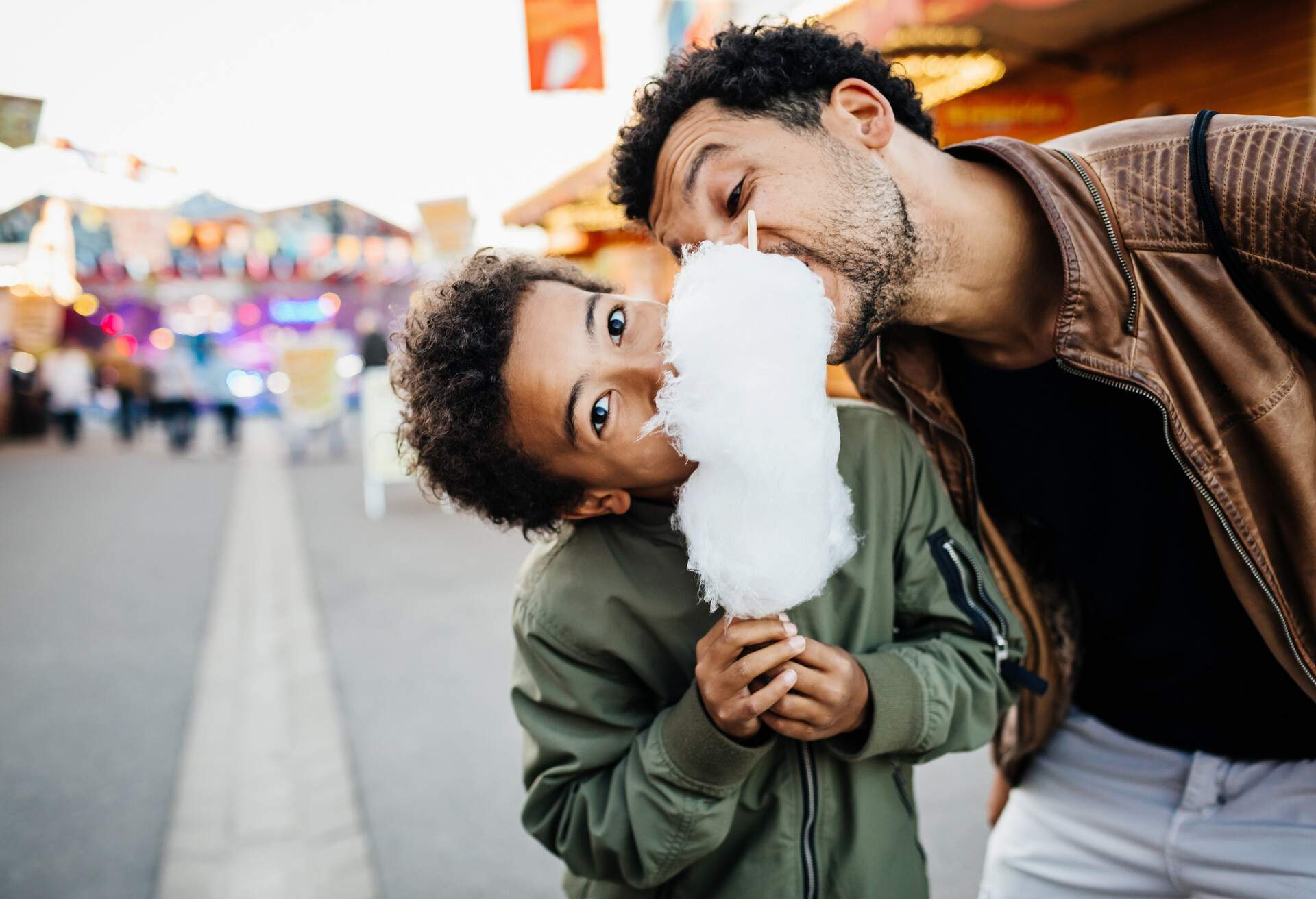 A playful father and son sharing a bloom of candy floss while spending the day at the fun fair together.