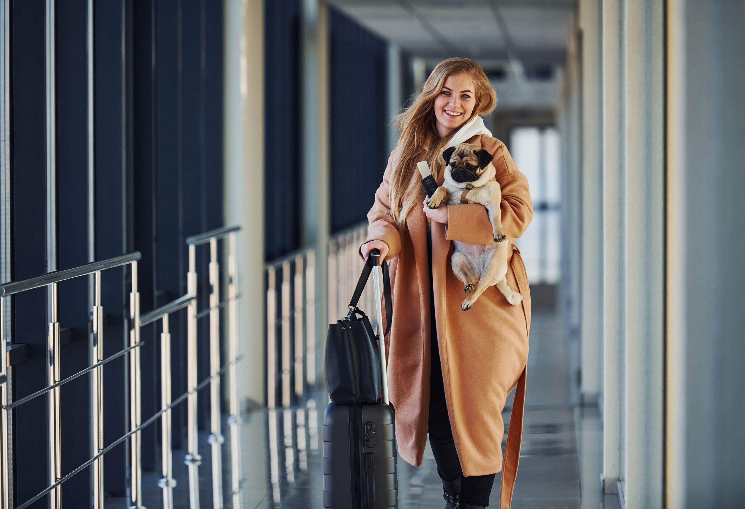 A smiling blonde woman holds her suitcase in one hand and carries her dog in the other.