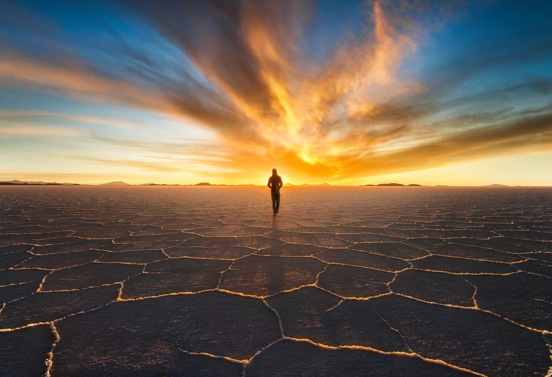 A person walks across a salt landscape with hexagonal patterns towards a sunset on the horizon.
