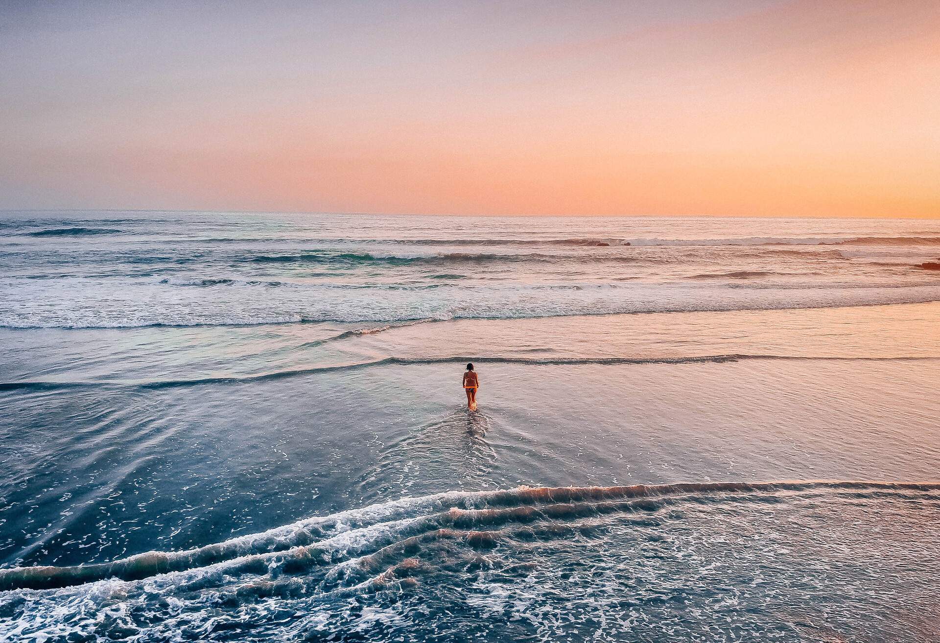 A person stands on a beach with calm waves under an orange sky.