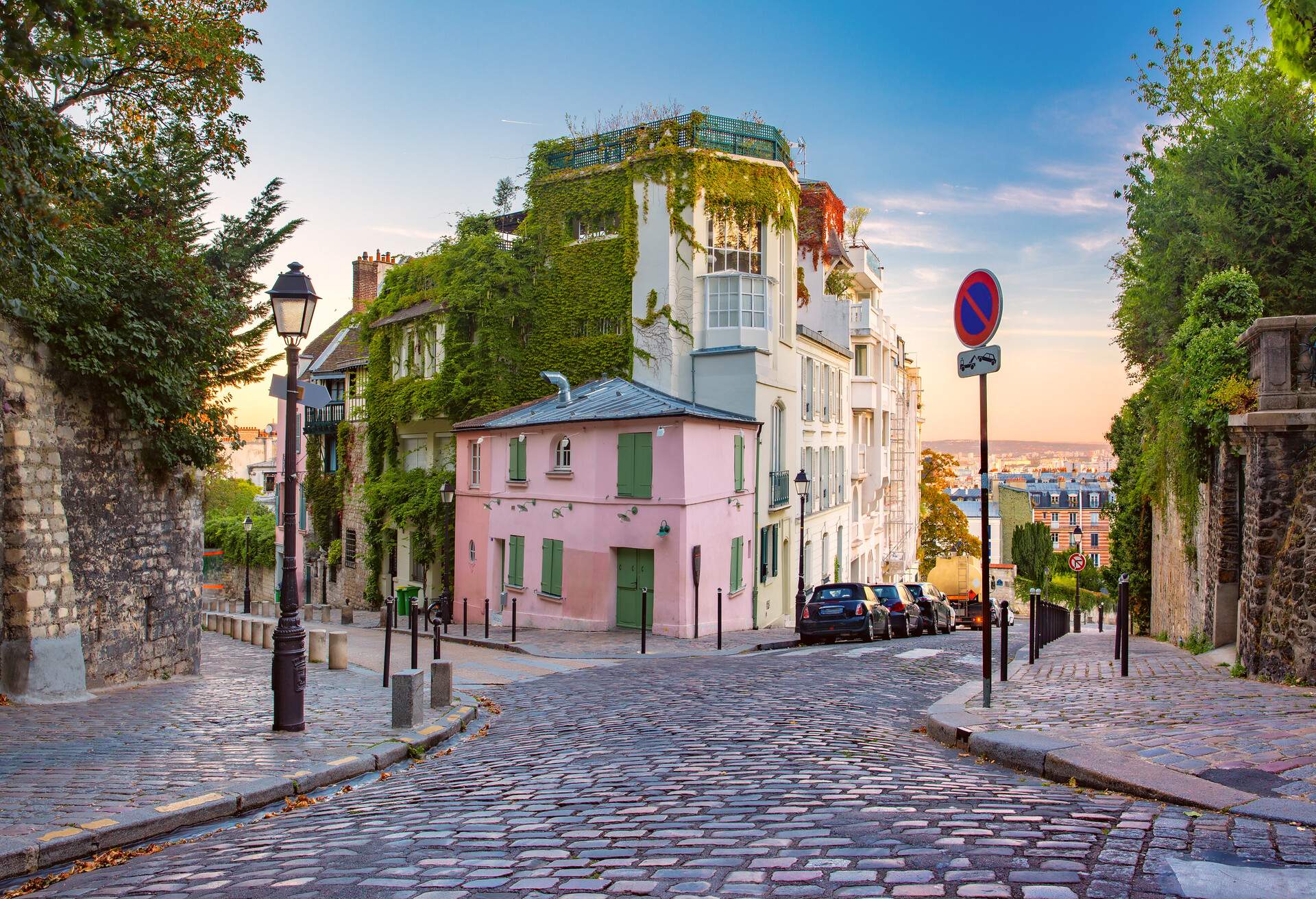 Charming quiet cobblestone street on a hilltop in Montmartre in Paris with a small pink house in the middle and a view of Paris in the background