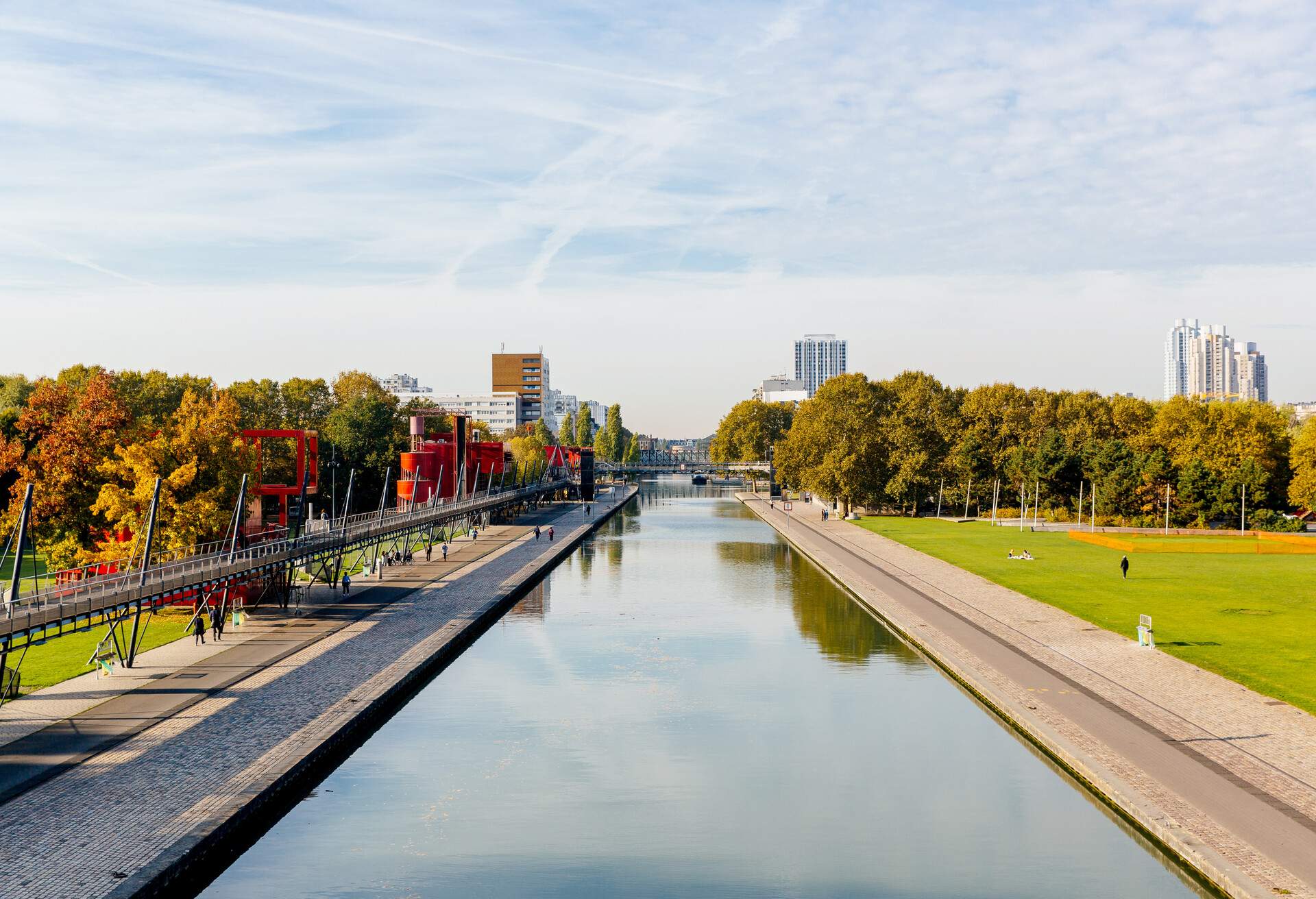 A canal in the middle of a park in Paris with tall industrial buildings peeping up in the horizon 