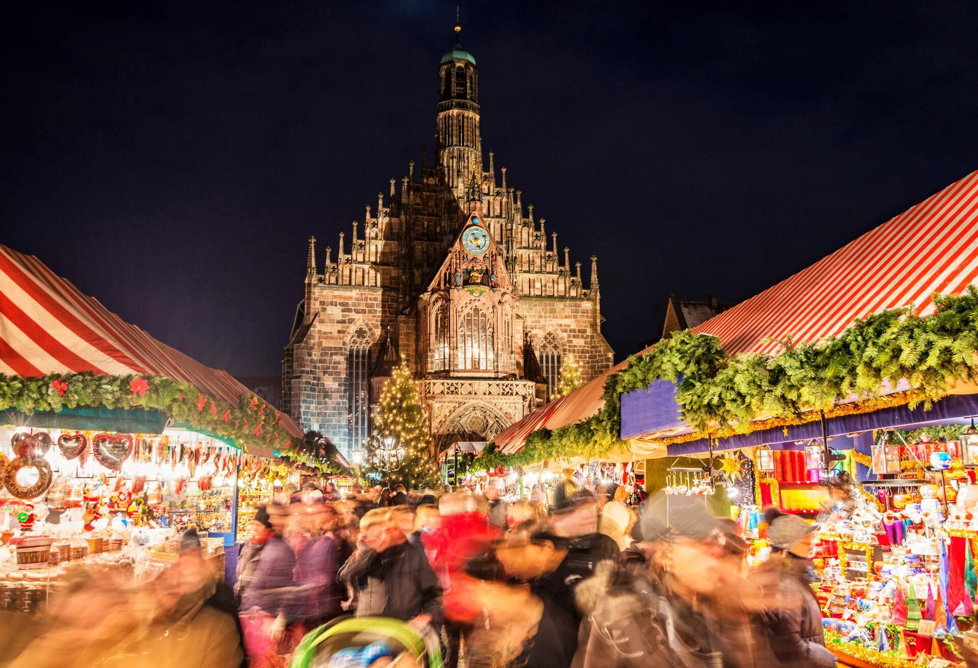 Huge crowd of people moving over Nuremberg´s world-famous christmas market (Christkindlsmarkt) at night, passing colorful illuminated christmas decoration and food stalls. Nuremberg´s landmark Frauenkirche (Church of our Lady) can be seen in the back.