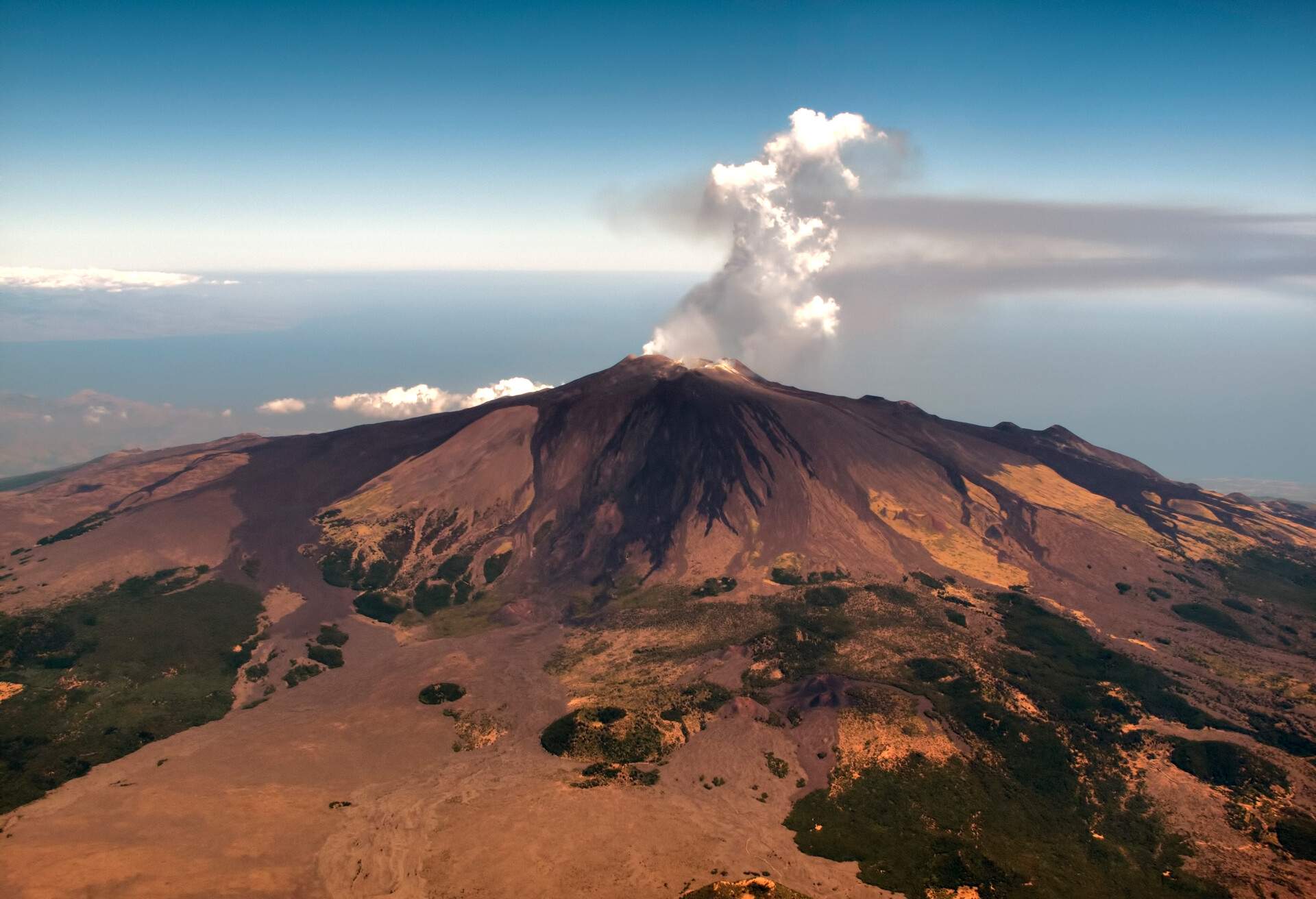 Aerial view of volcanic activity on top of Mount Etna, largest and most active volcano in Europe, with central crater spewing ash and smoke on  clear blue sky.