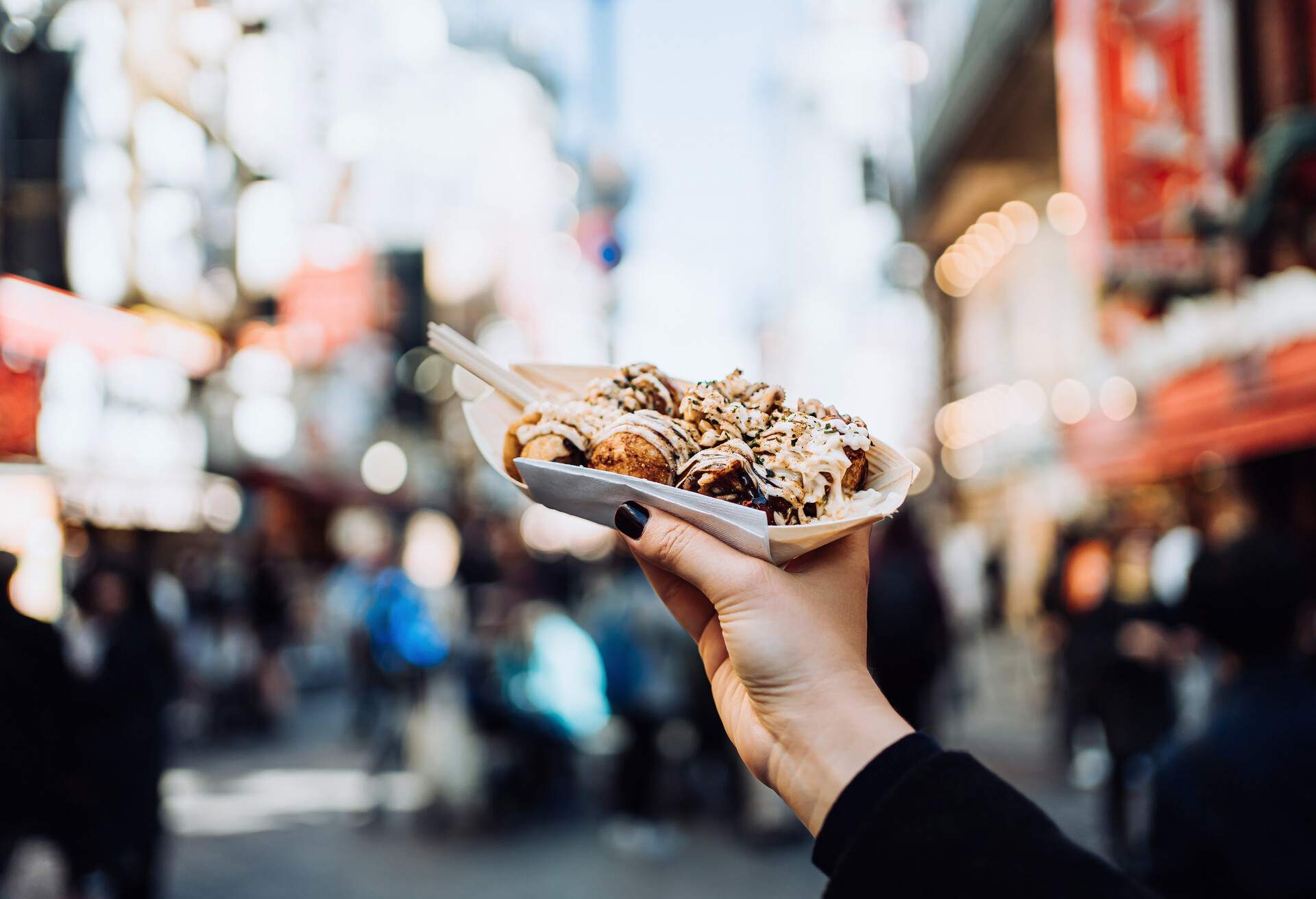 Personal perspective of a female traveller holding freshly made traditional Japanese street-style snack takoyaki (octopus balls) against downtown city street while visiting Osaka
