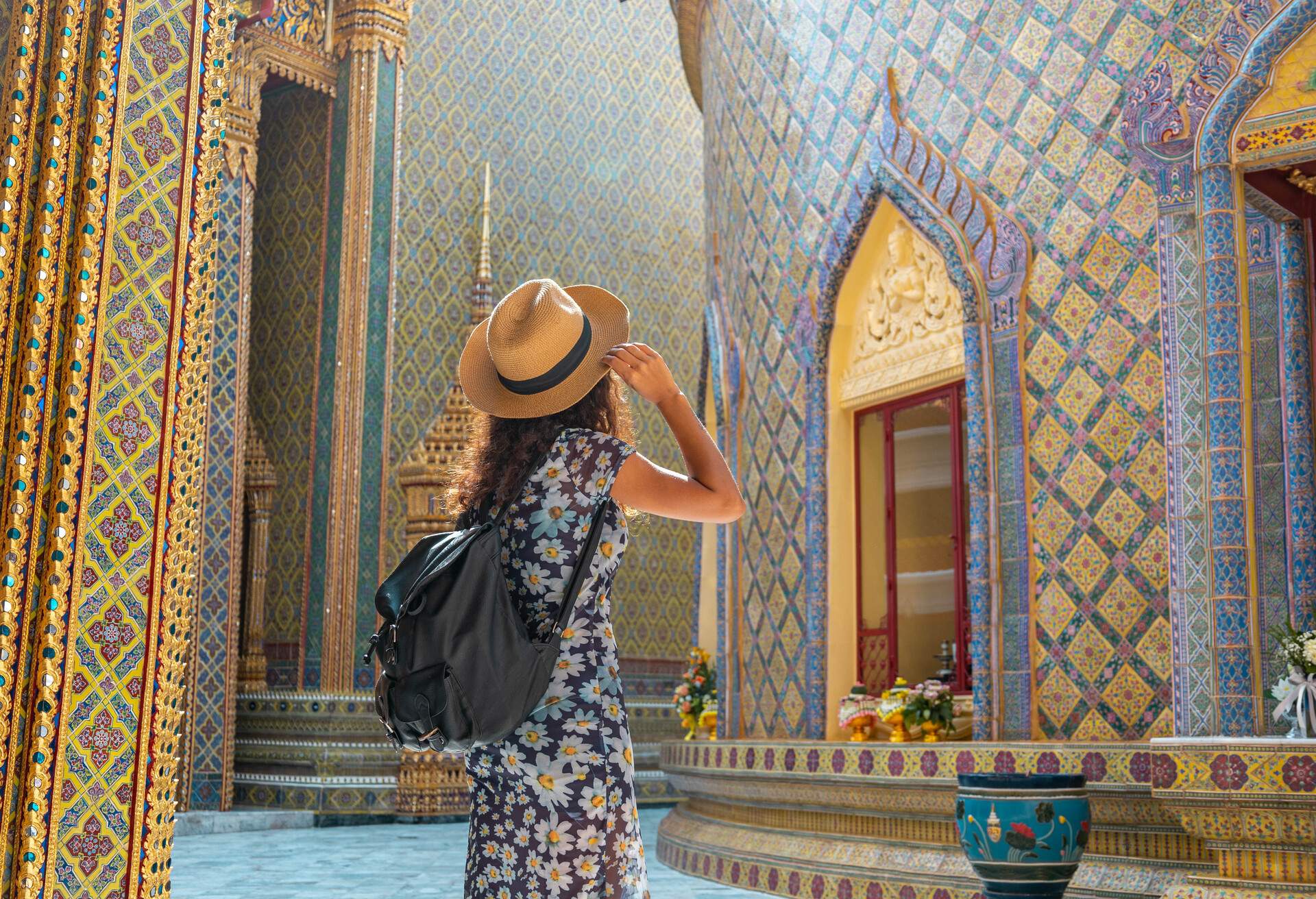 Woman in a floral dress touches her hat and looks up at the intricately crafted ceramic and golden walls of a temple.