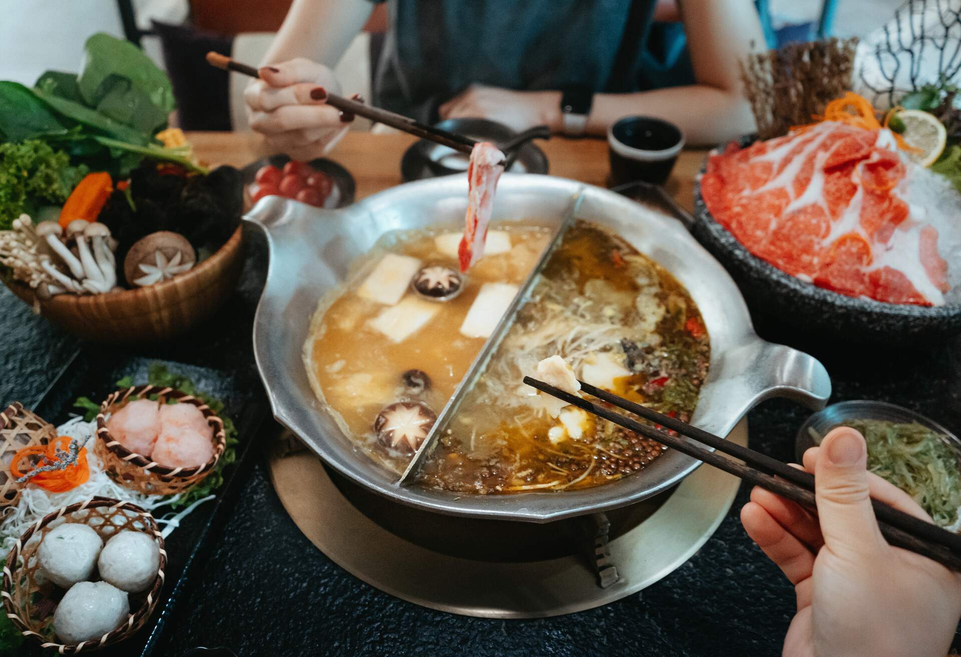 Personal perspective of Asian couple having a good time, enjoying traditional Chinese hotpot with assorted fresh and scrumptious ingredients in restaurant. Chinese cuisine and food. Eating out lifestyle