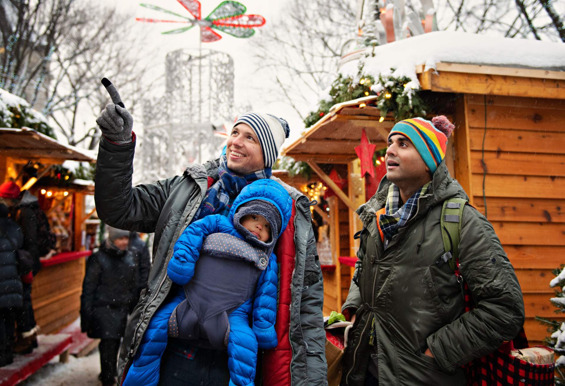 Married gay fathers shopping for Christmas with their one year old mixed race  baby adopted at the Christmas Market in Quebec Canada. One of the daddy carrying his baby in a backpack in front of him. The guide follow them to explain the ancestral life of Quebec.