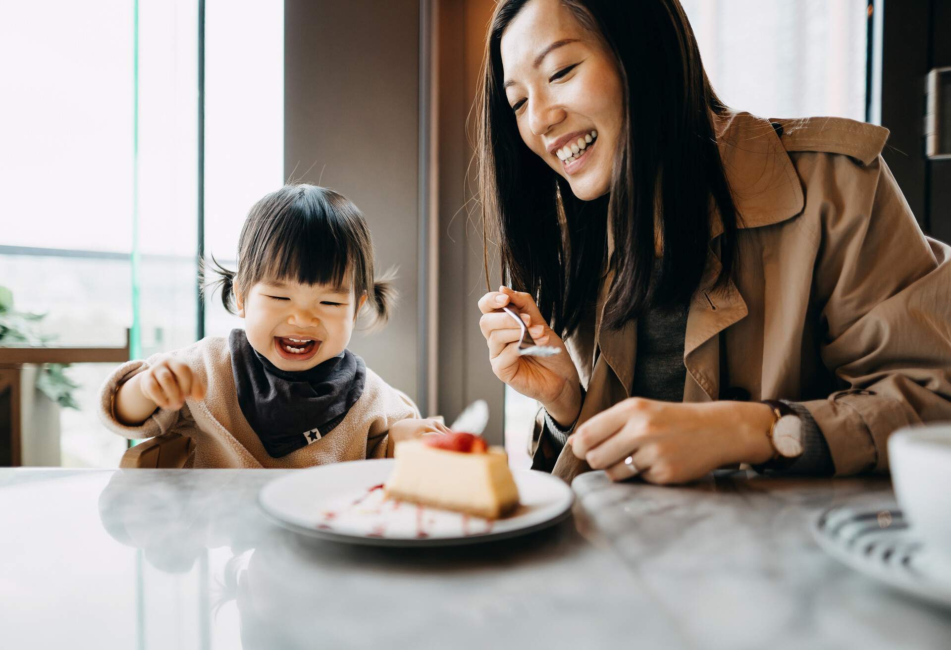 Mother and little daughter sharing a piece of cake in the cafe, both of them are smiling joyfully