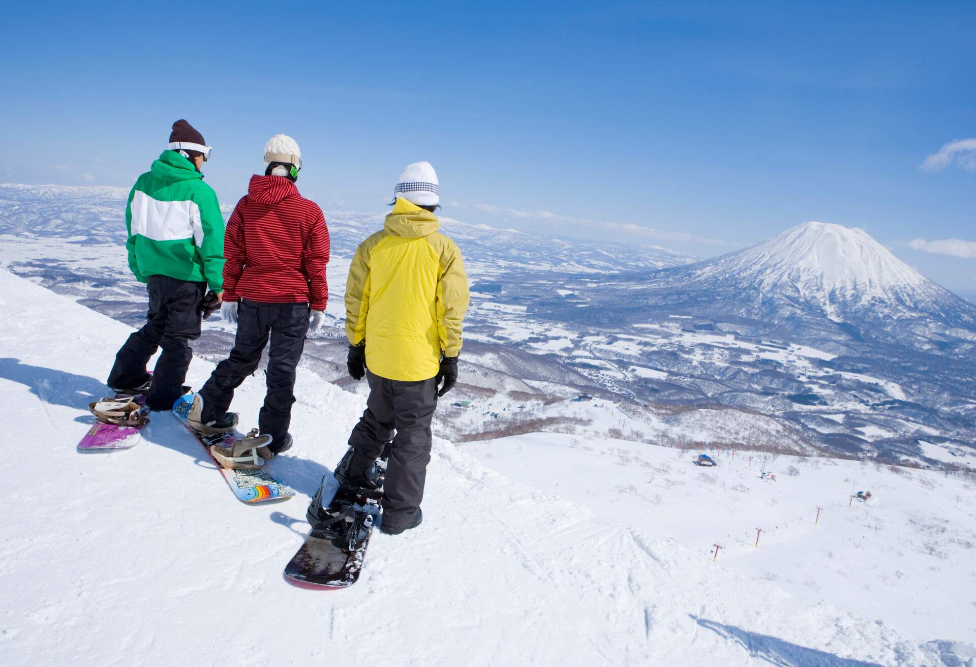 Three male friends in winter clothes stand on their snowboards on a snow-covered mountain looking at the ice-capped coned shape volcano below.