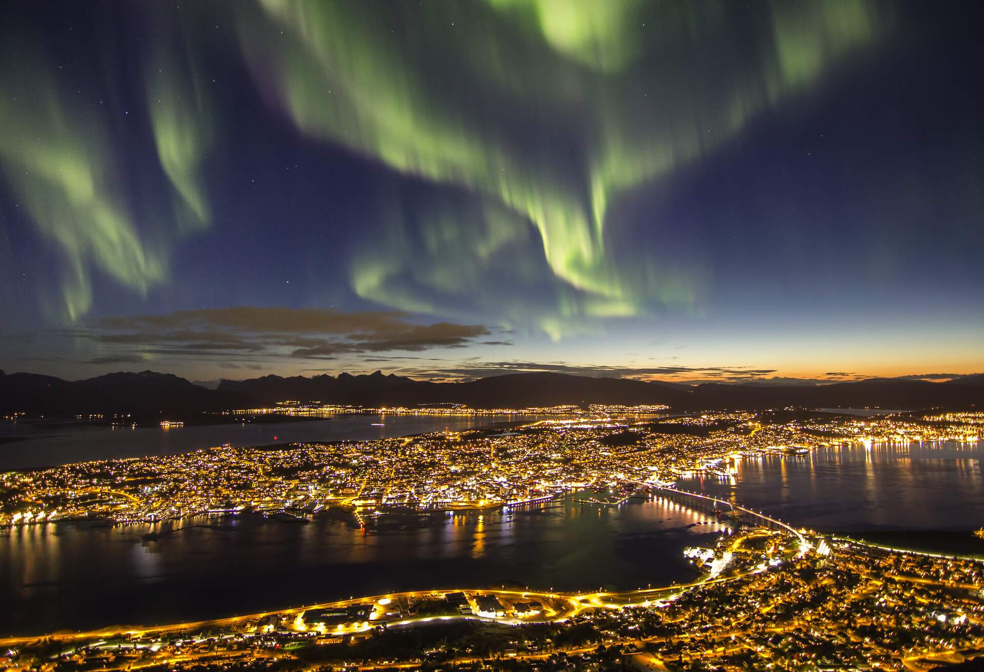 A night-time view of a dazzling coastal city under the green streaks of northern lights.