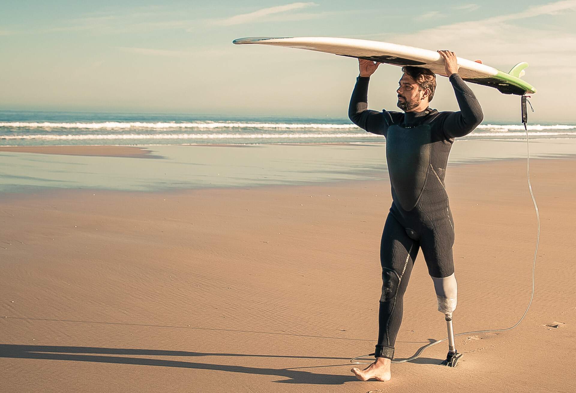 Male surfer wearing artificial limb and wetsuit, walking on beach, carrying surfboard overhead. Full length. Artificial limb and active lifestyle concept