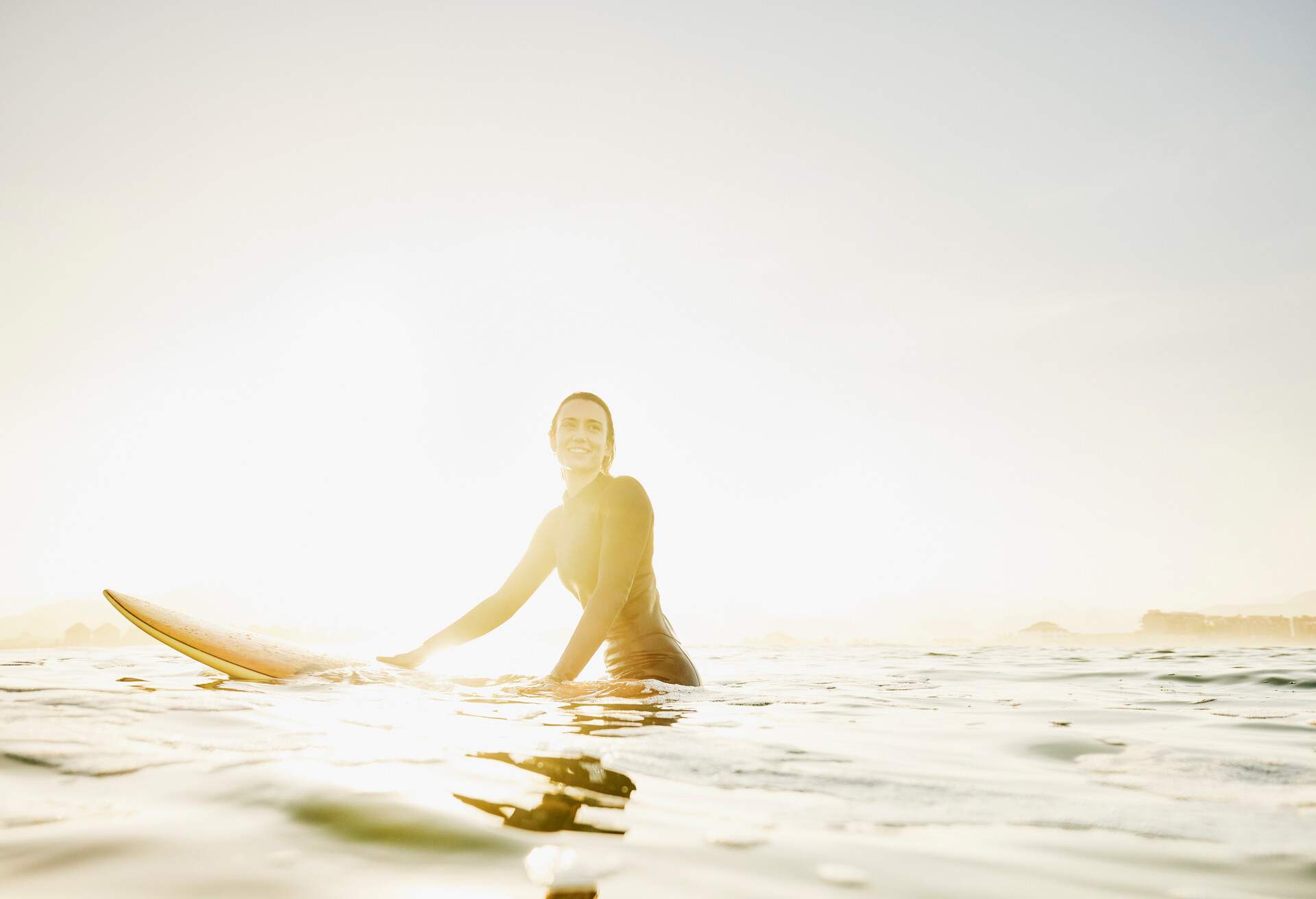Wide shot portrait of smiling female surfer sitting on surfboard in ocean while waiting for wave during early morning surf session