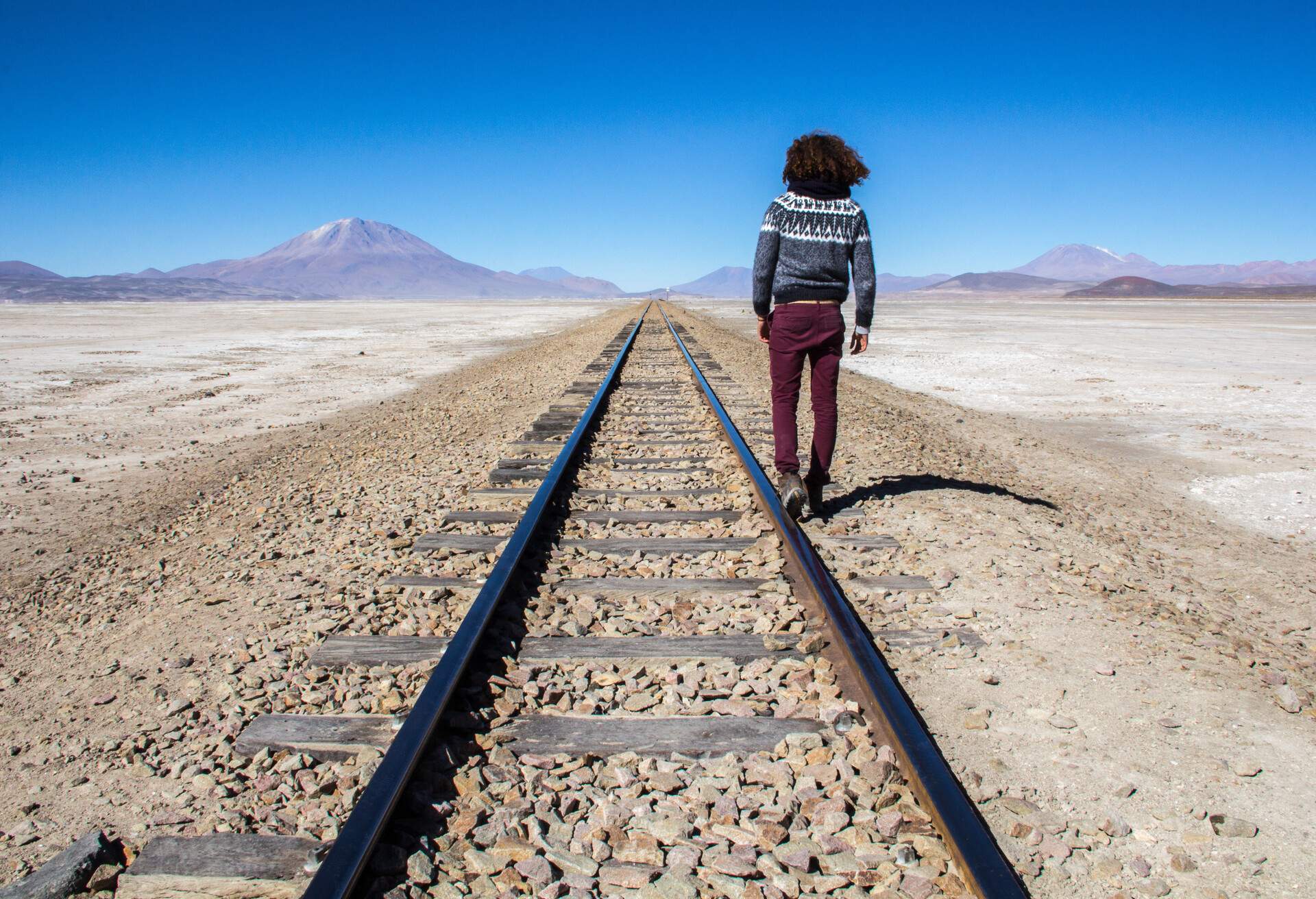 Potosi, Bolivia- October 30, 2016: Man with Afro, wearing a grey sweater, brown pants and boots, walking on railroads that seems to never end. The sky is really blue and there are some mountains