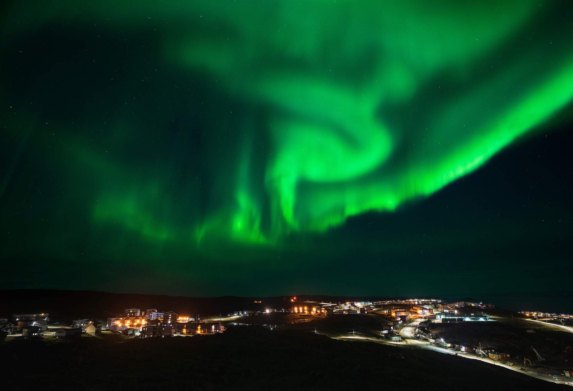 Northern lights with a swirling green tint hovering above the city's lit streets and surrounding structures at night.