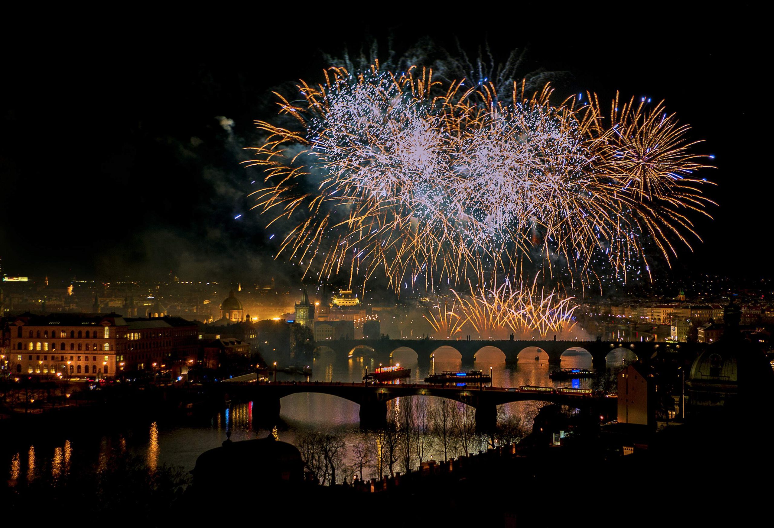 Exploding fireworks lit the dark sky above a broad river spanned by bridges in a populous city.