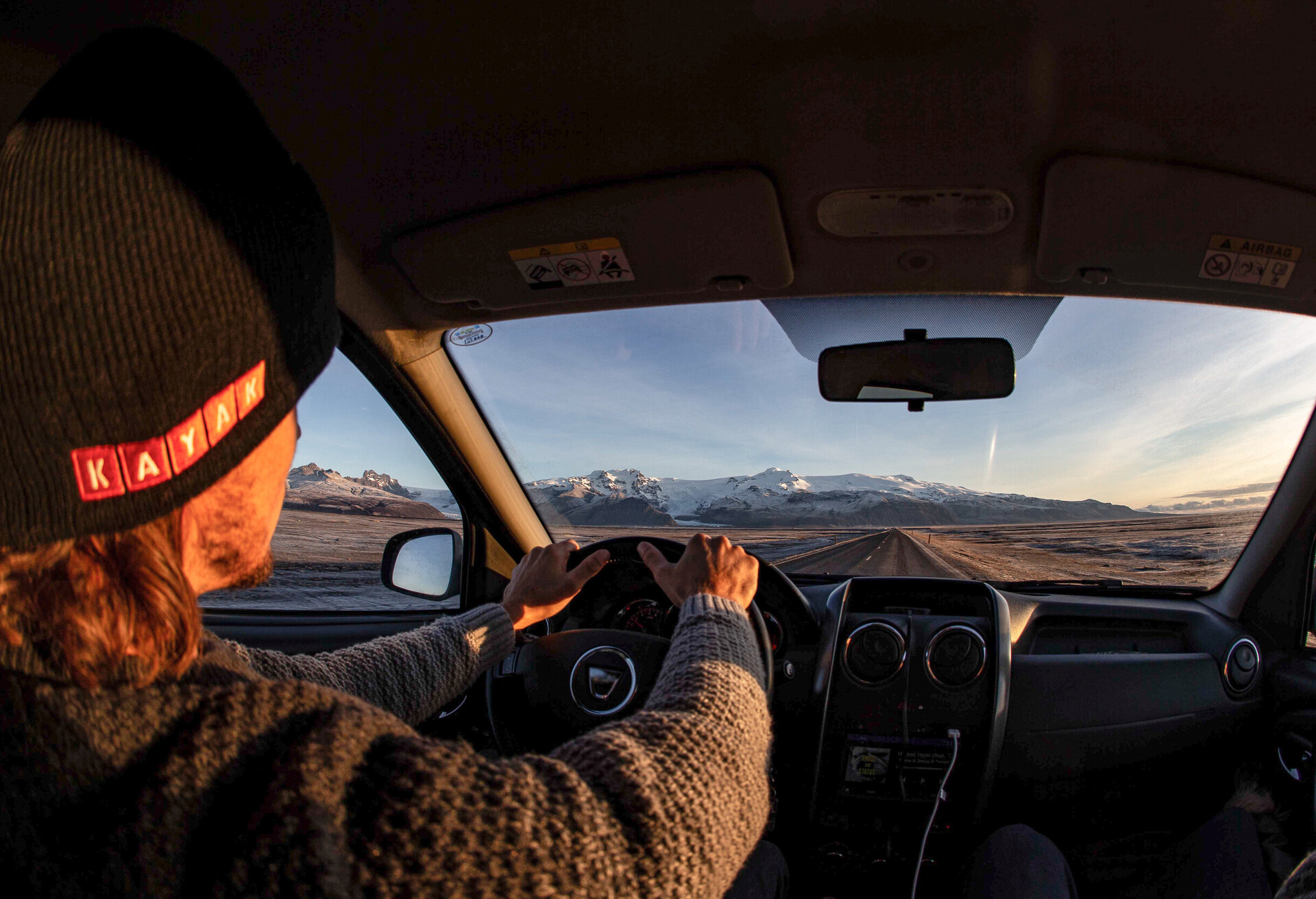A driver holding the car's steering wheel with views of an empty road and snow-capped mountains.
