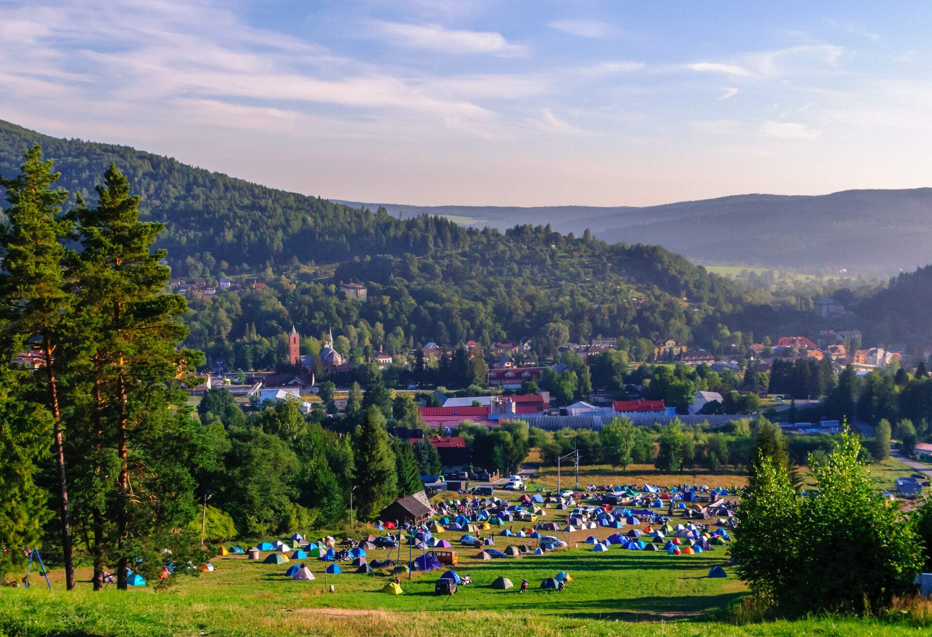 A cluster of tents on a campsite bordered by trees, with a town in the distance.