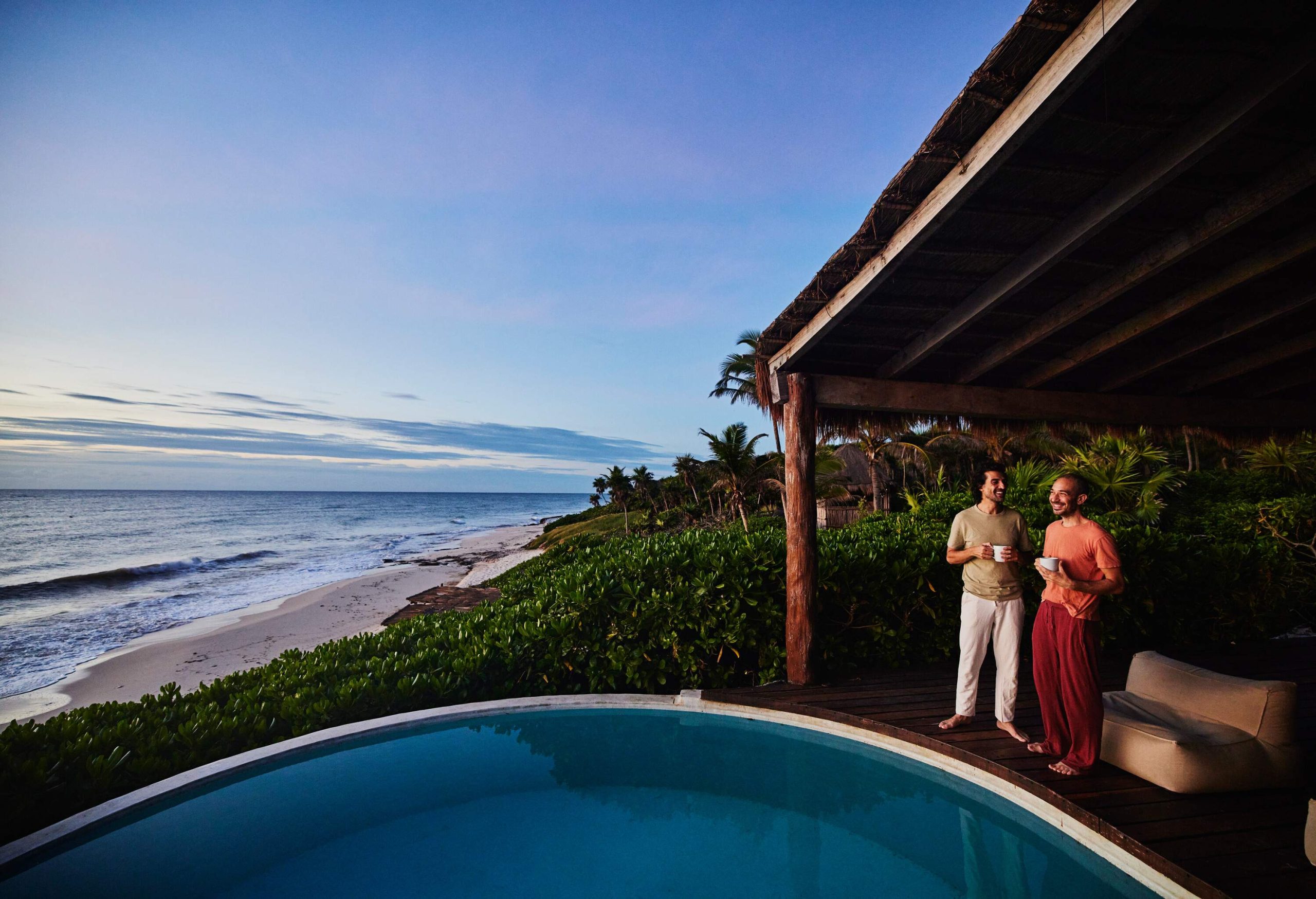 Two men holding white cups and standing by a pool overlooking the beach.