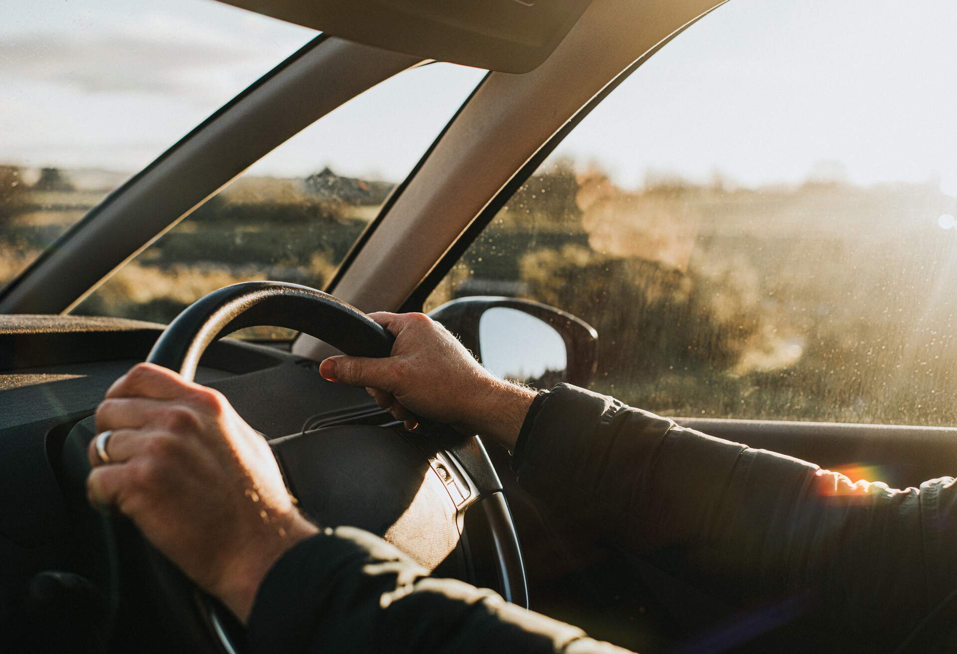 Interior of a car. Hand holds a black steering wheel. Car is illuminated by low, warm sun at dusk.