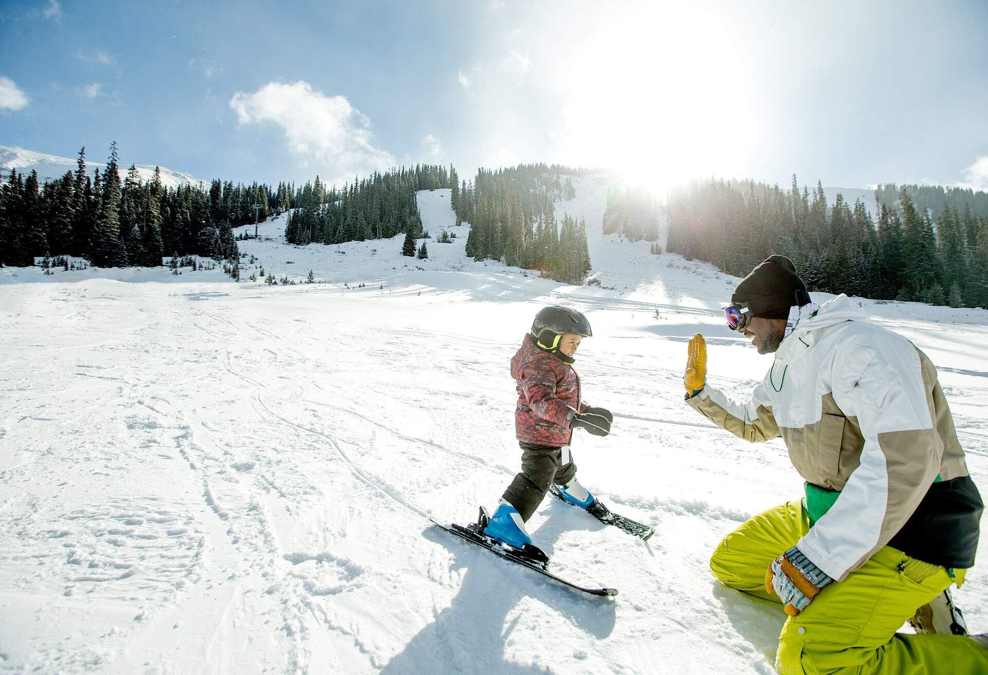 A man dressed for the winter raises his hand to high-five a young skier on a snow field.