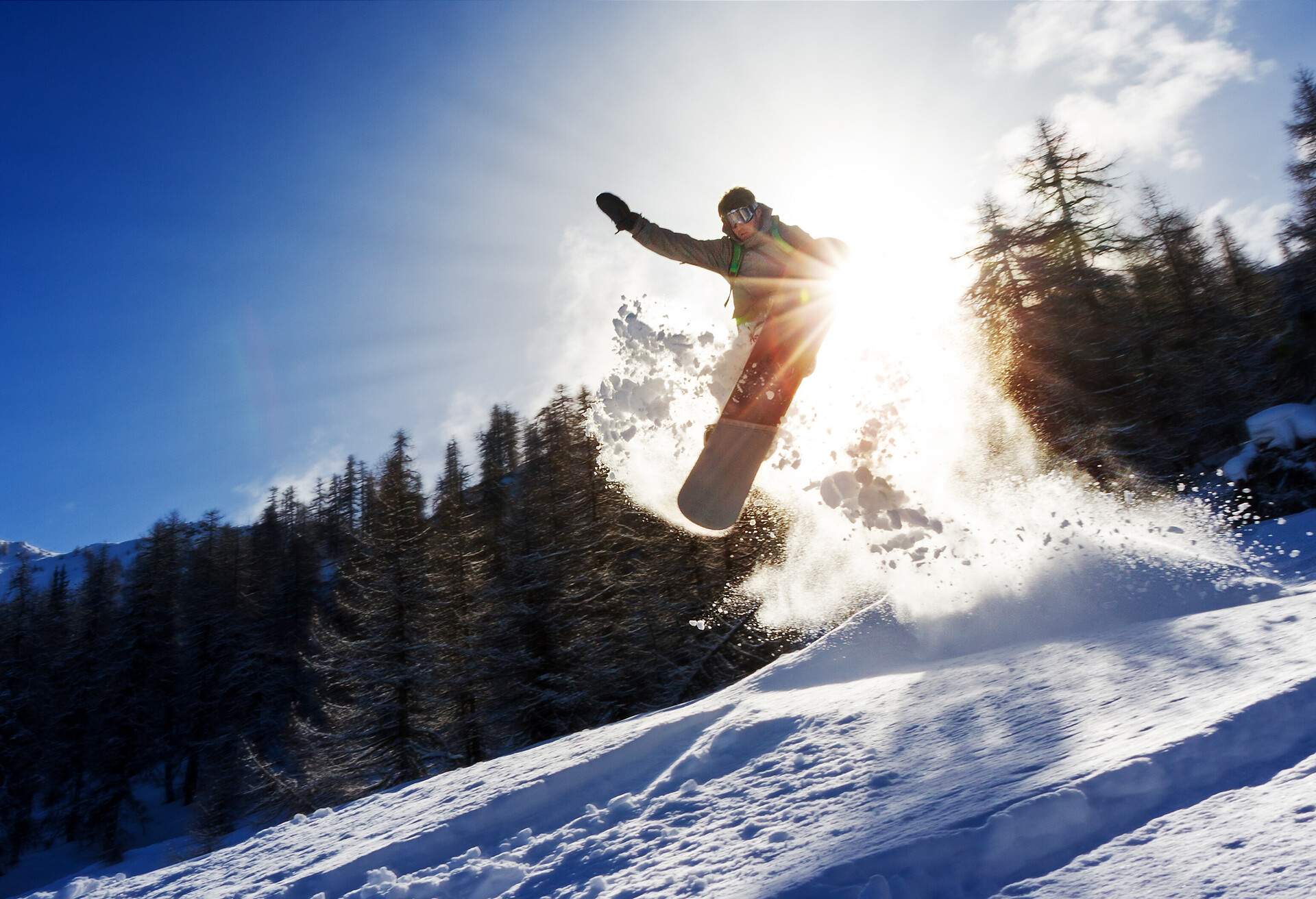 A snowboarder jumping on a snowy ski slope as the sun shines through the trees in the background.