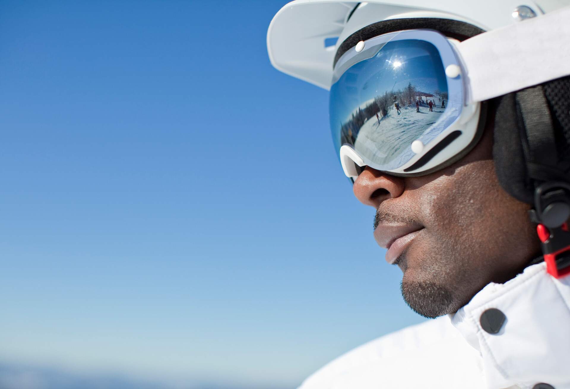 A man in white ski gear with ski landscape reflected on the lens of his goggles.