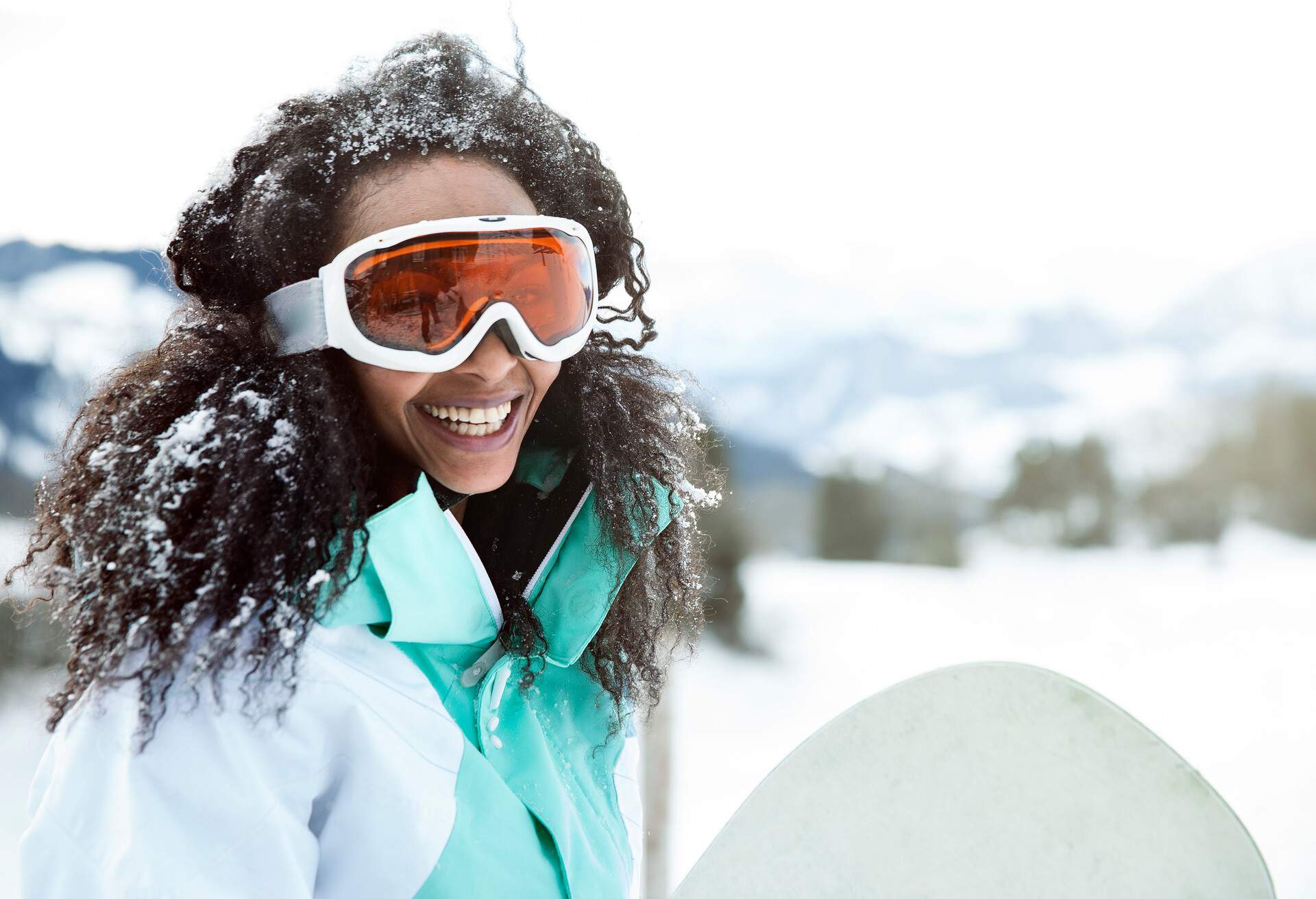 A cheerful young lady wearing white-framed goggles standing with a snowboard, hair peppered in snow. 