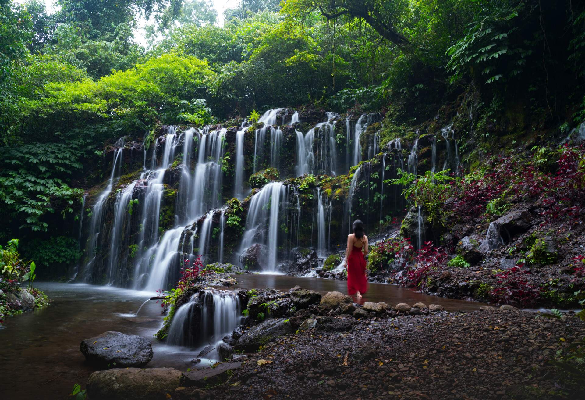 Young beautiful aisna girl in red skirt with tanned skin explore Banyu Wana Amertha Waterfall during her summer vacation in Bali, Asia 