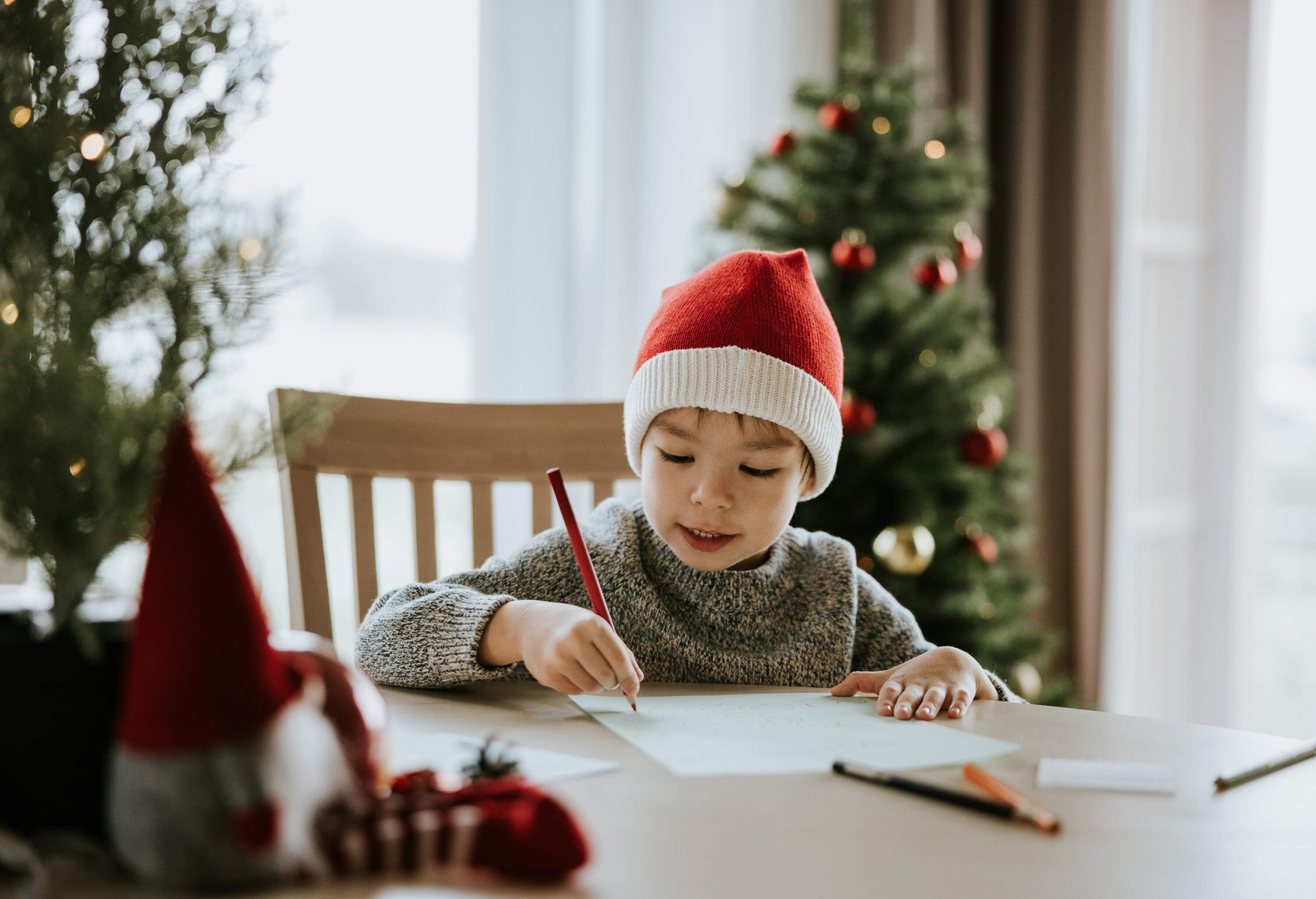 a little boy sitting by a table writing a letter wearing a santa hat