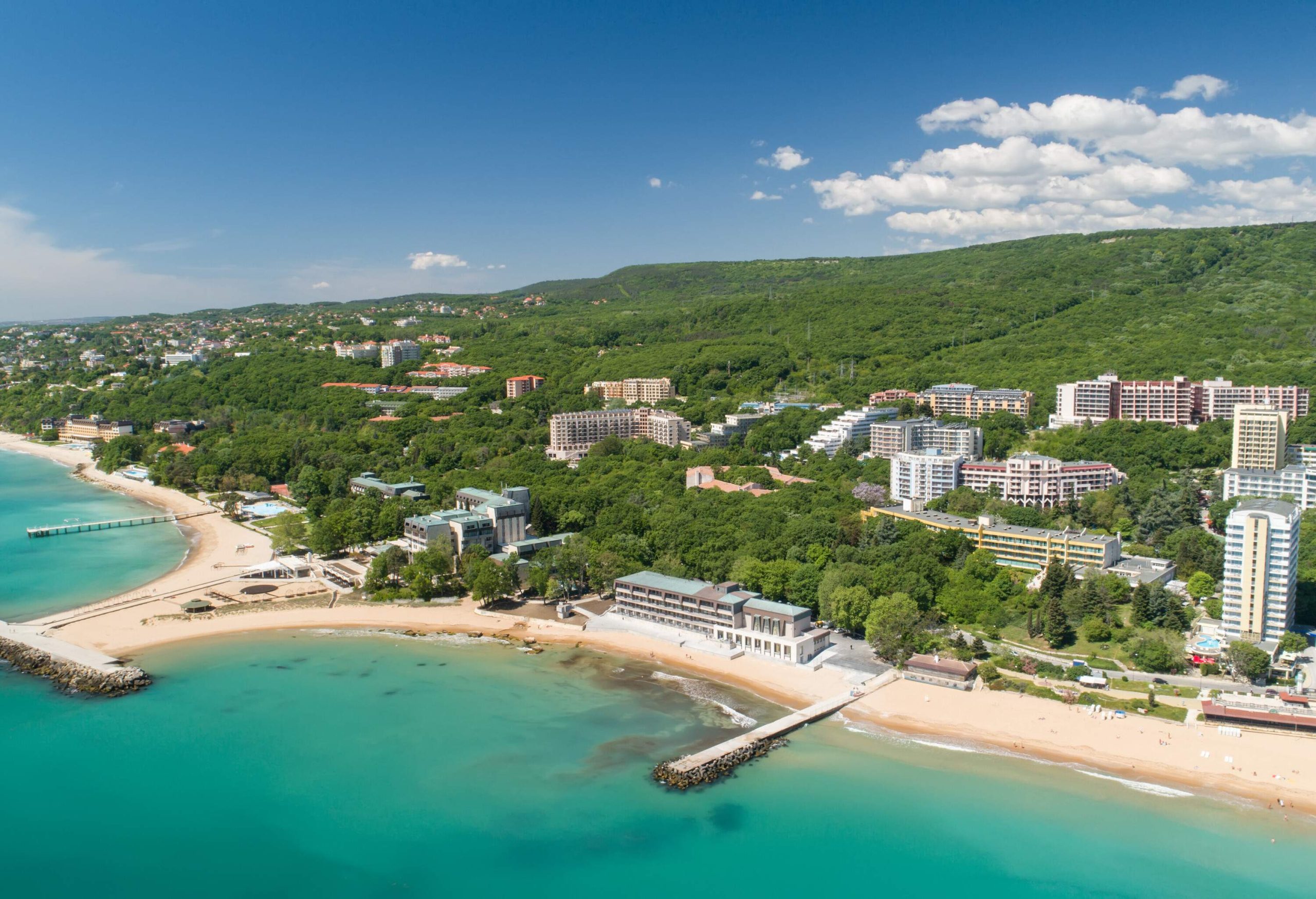 A beach with artificial breakwaters and a cluster of buildings perched on forested hills.
