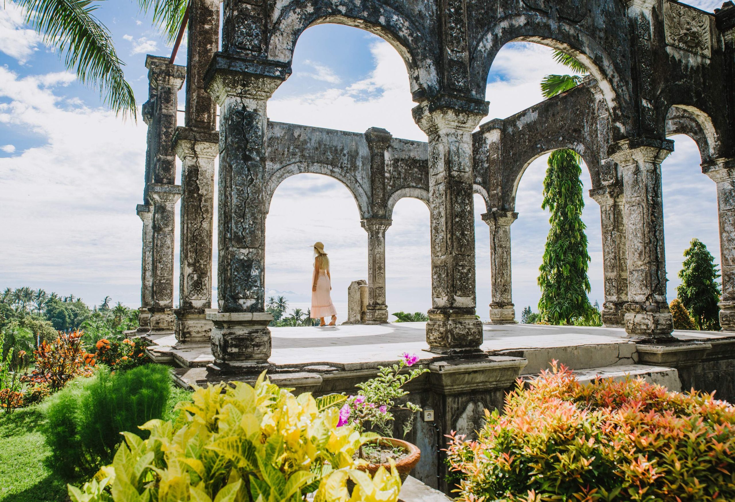 A woman stands in the middle of the ruins of a temple with an open roof and arched columns, surrounded by lush landscapes and colourful flowers.