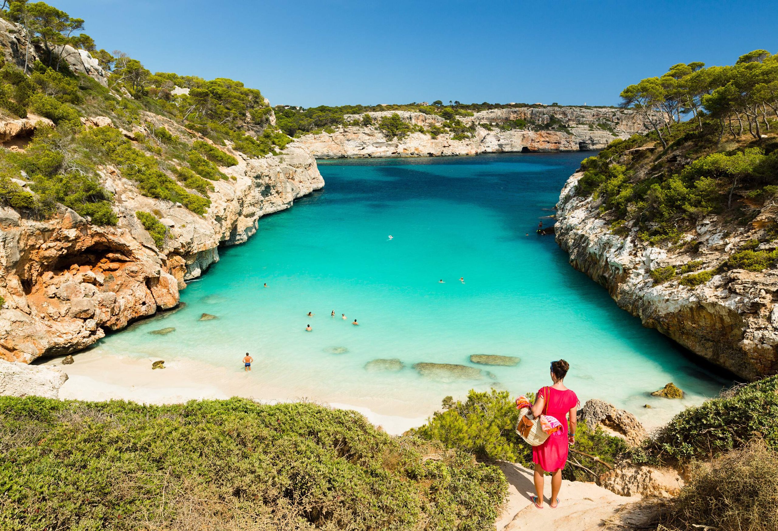 A woman in a pink dress looks at her friends down the beach with white sand and clear waters. 