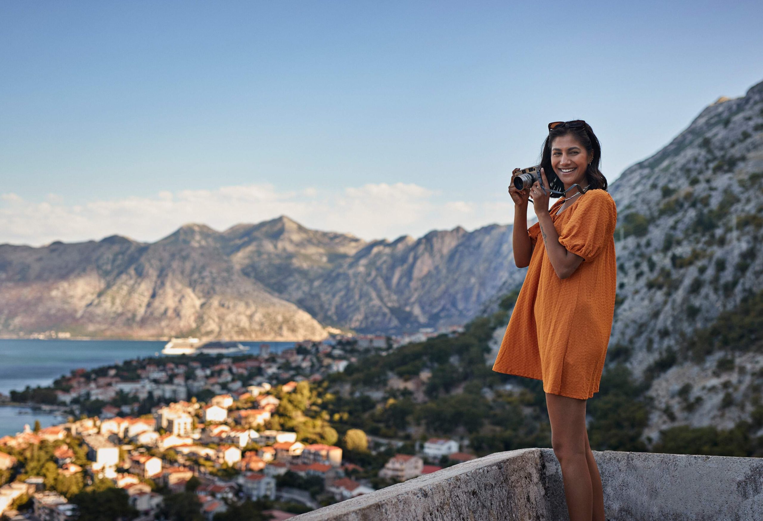 A woman in an orange dress smiling while holding a camera against a backdrop of a seaside town.