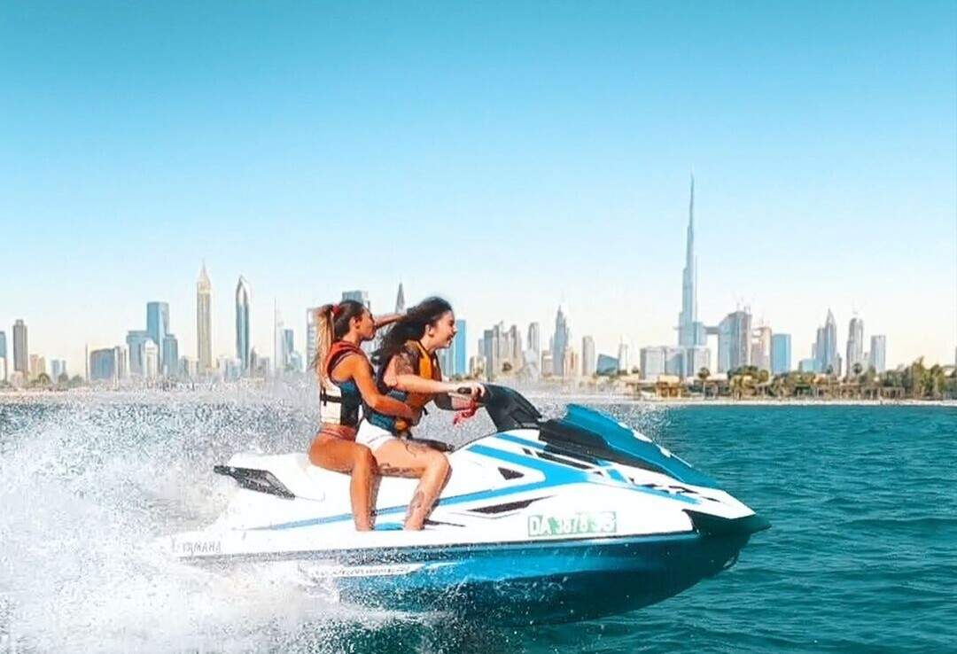 Two ladies fastly drive a jet ski on green waters with a cityscape in the distance.