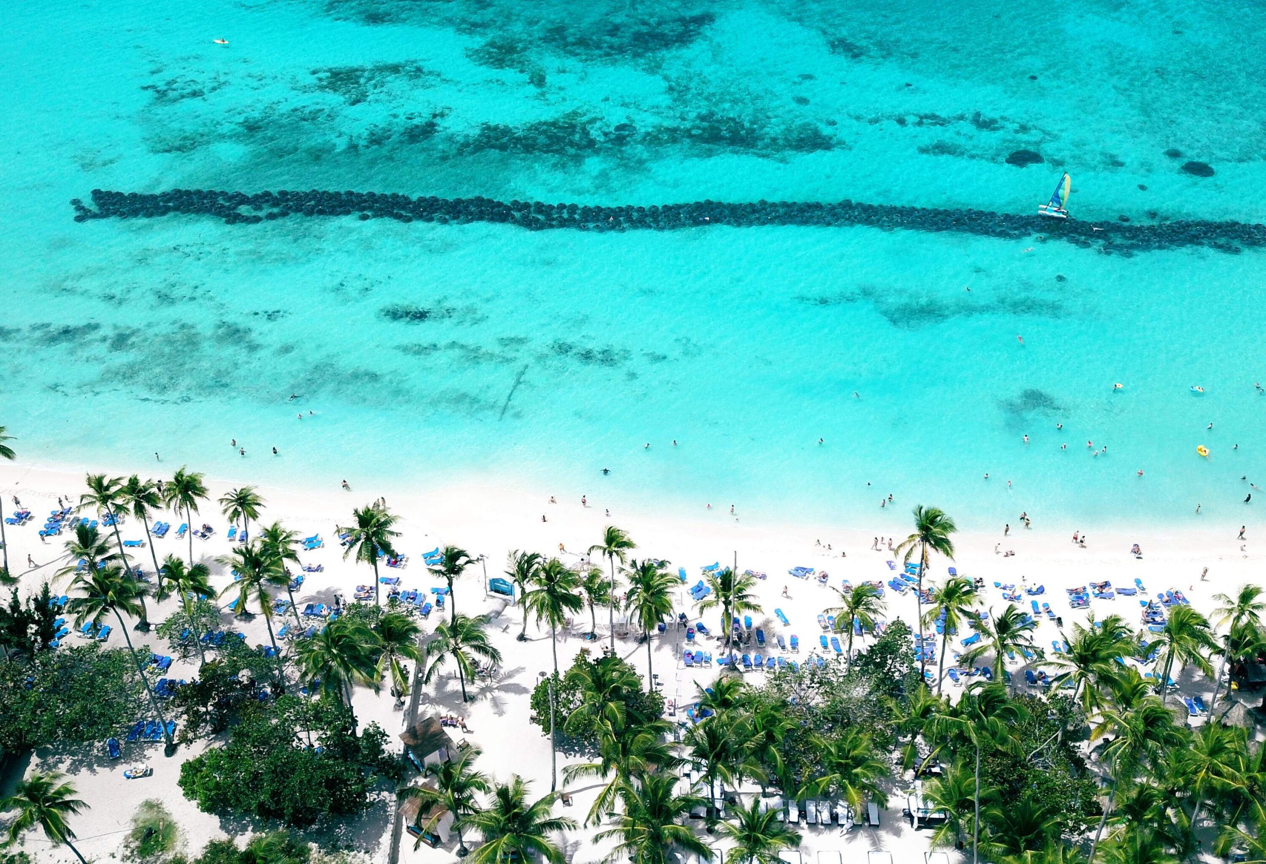 A crowded white sand beach with a cluster of sunbeds along a grove of palm trees beside the crystal-clear turquoise waters.