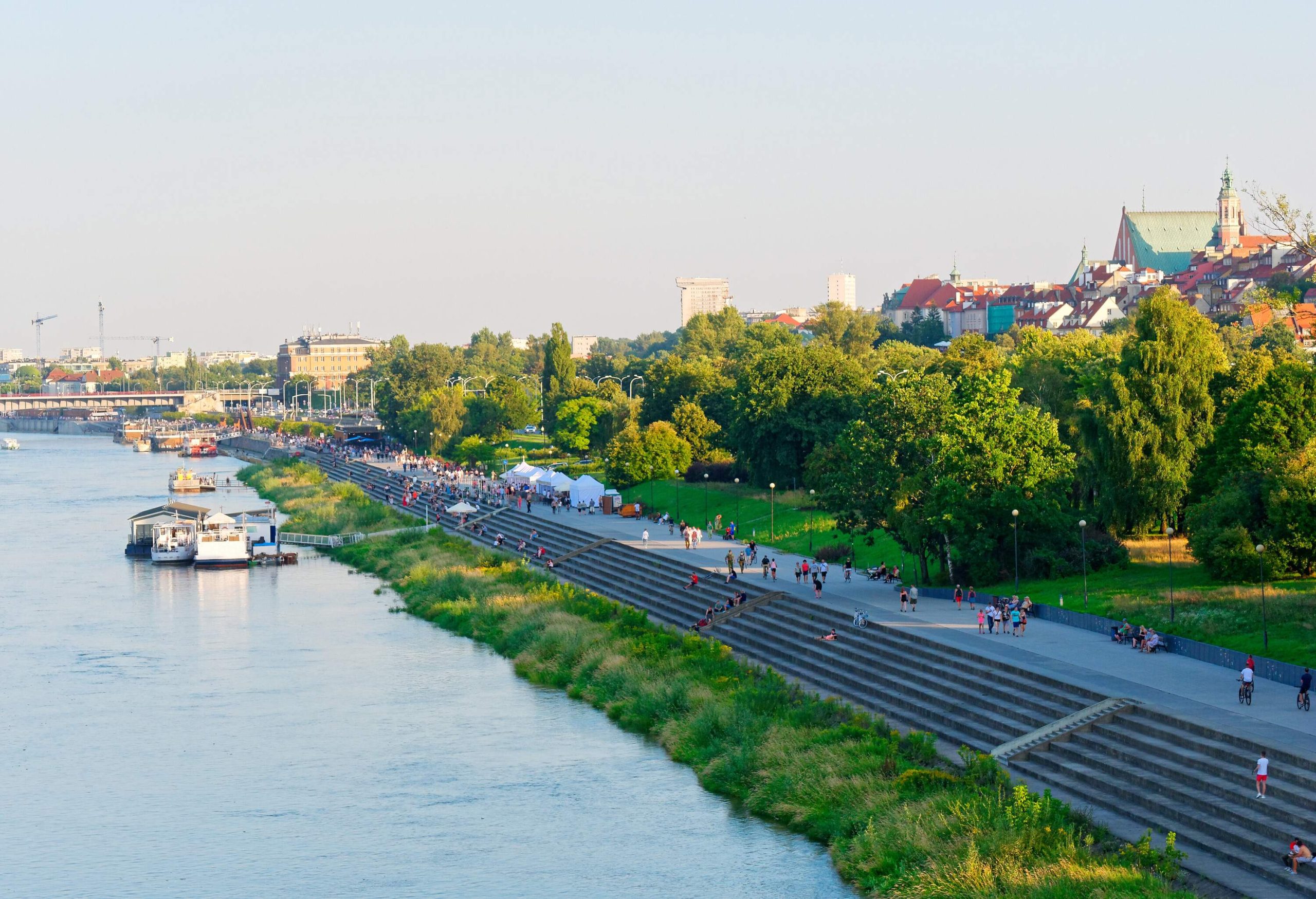A promenade on a lush green riverbank with stairs descending to the water with several boats and cafes on the tents.