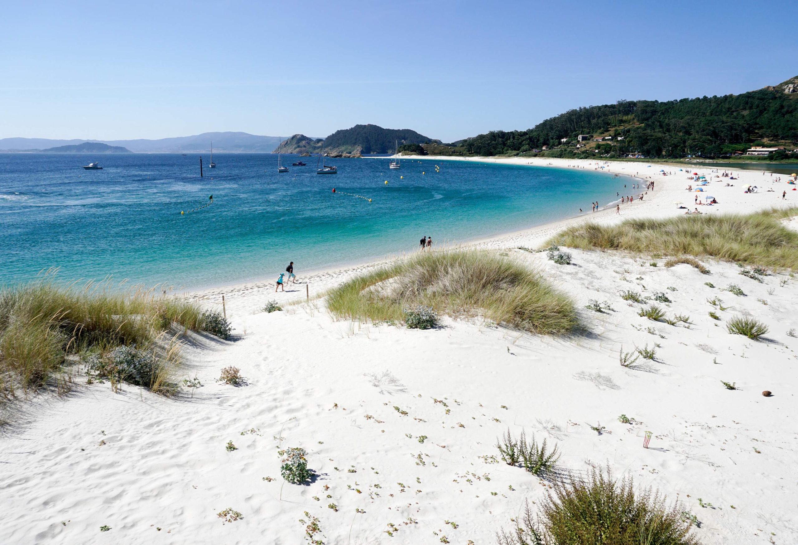 A white sand beach with boats and people is seen at the base of a lush, forested hill.