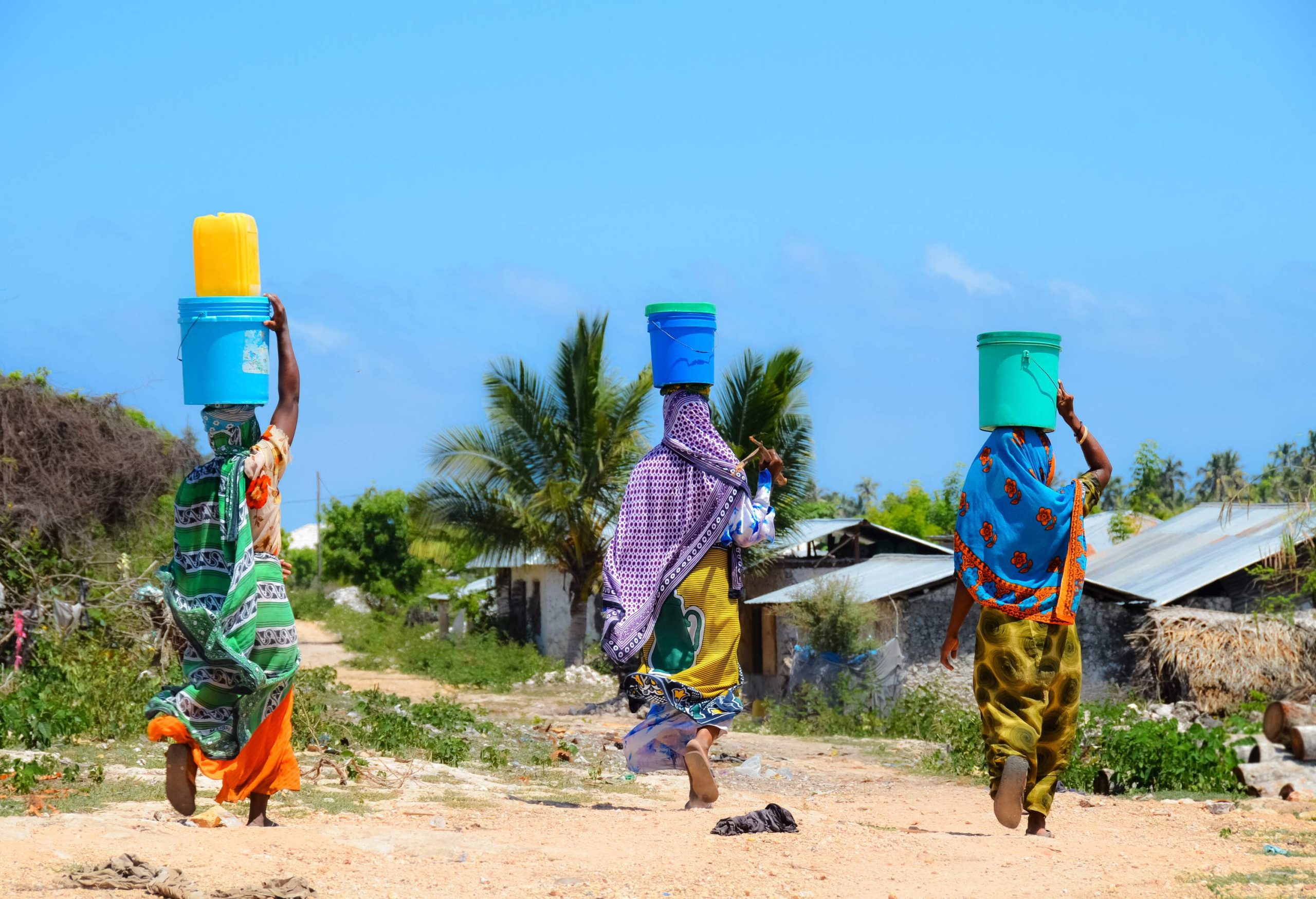 A rear view of three women carrying buckets on their heads walks toward the houses.