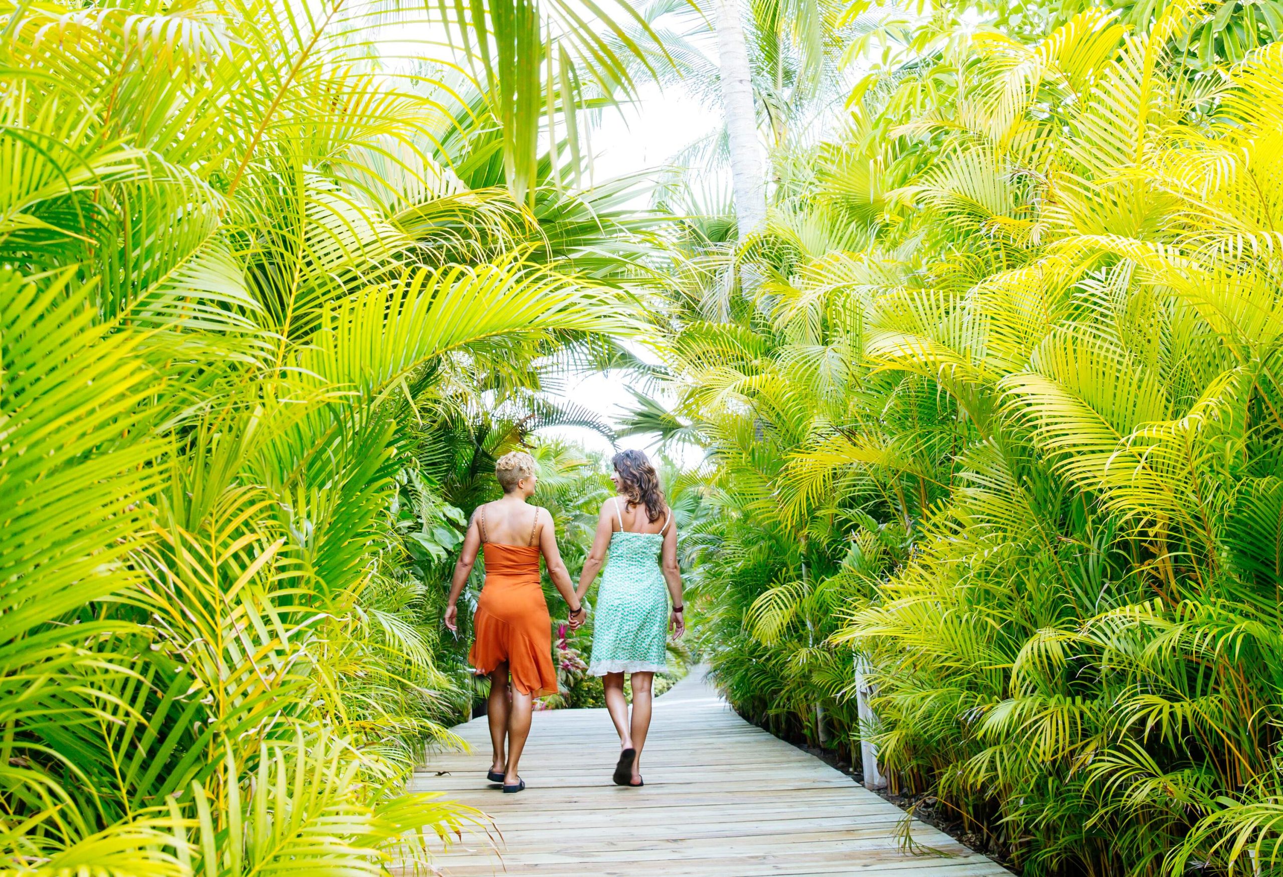 Two women holding hands while walking on a wooden footpath lined with palm trees.