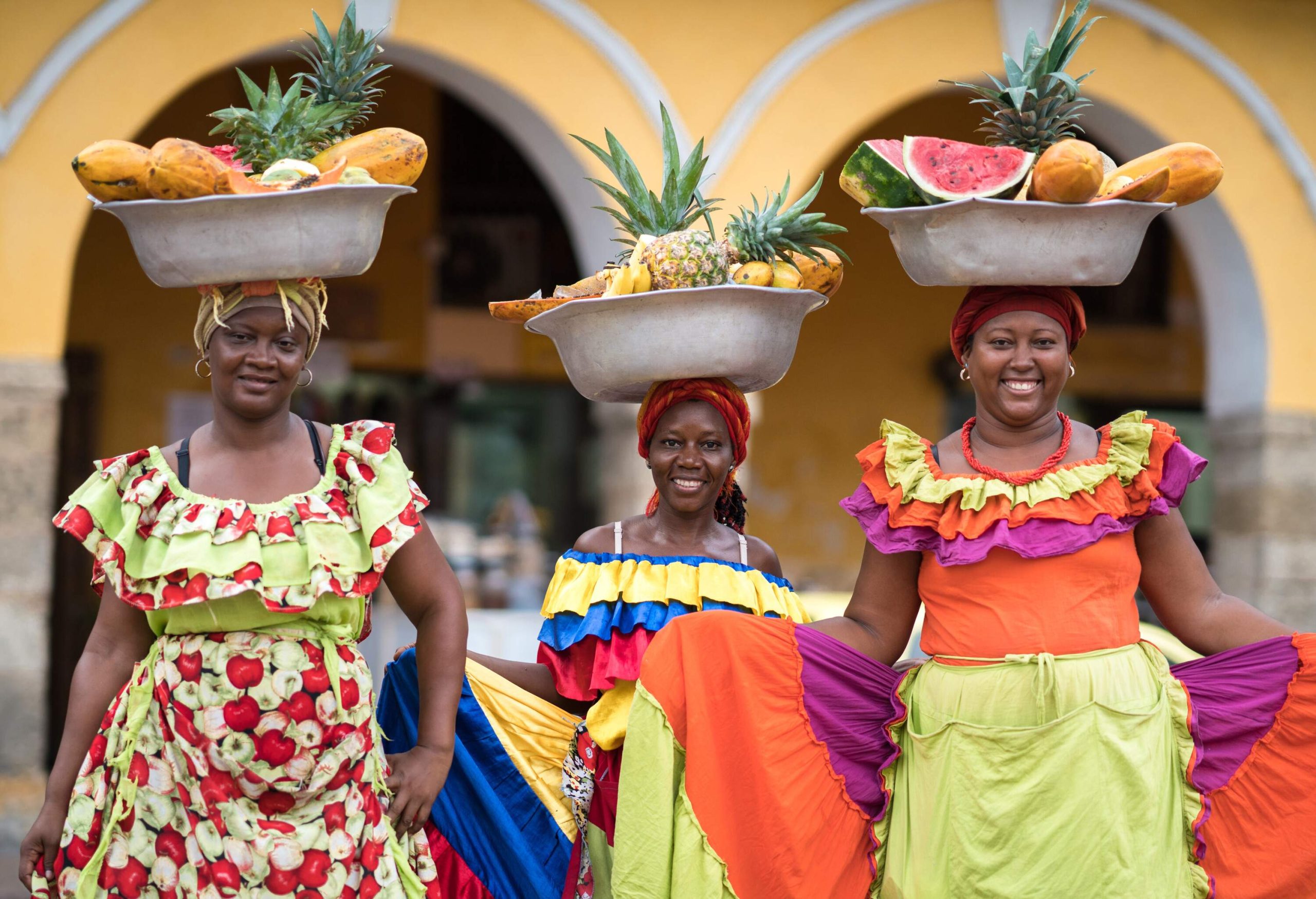 Three joyful women, dressed in colourful attire, skillfully balancing basins of fruits on their heads.