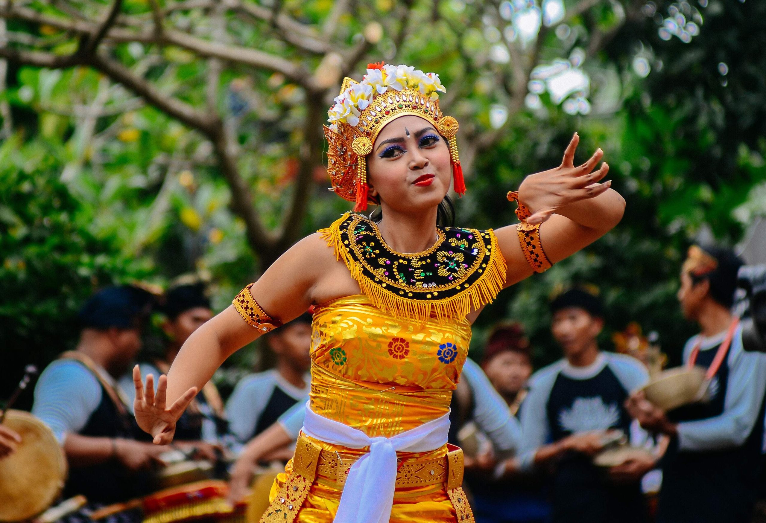 A woman dancing wearing a traditional Balinese costume, accompanied by a band playing behind her.